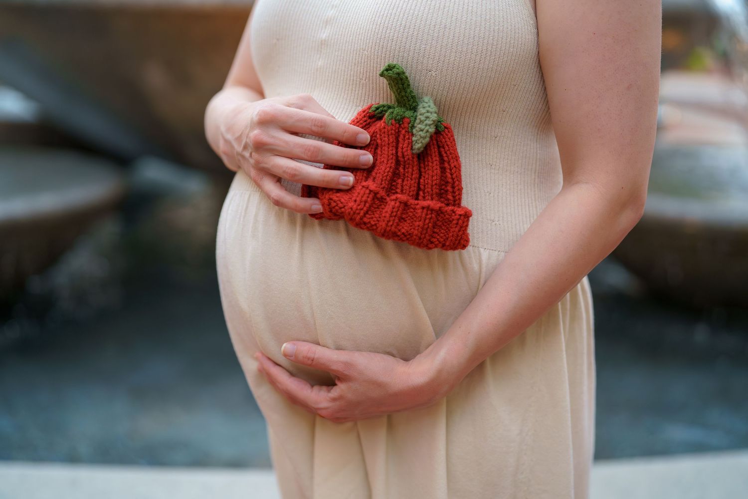 A woman in a peach-colored dress holds a red knitted baby bootie against their pregnant belly, in Alexandria, VA