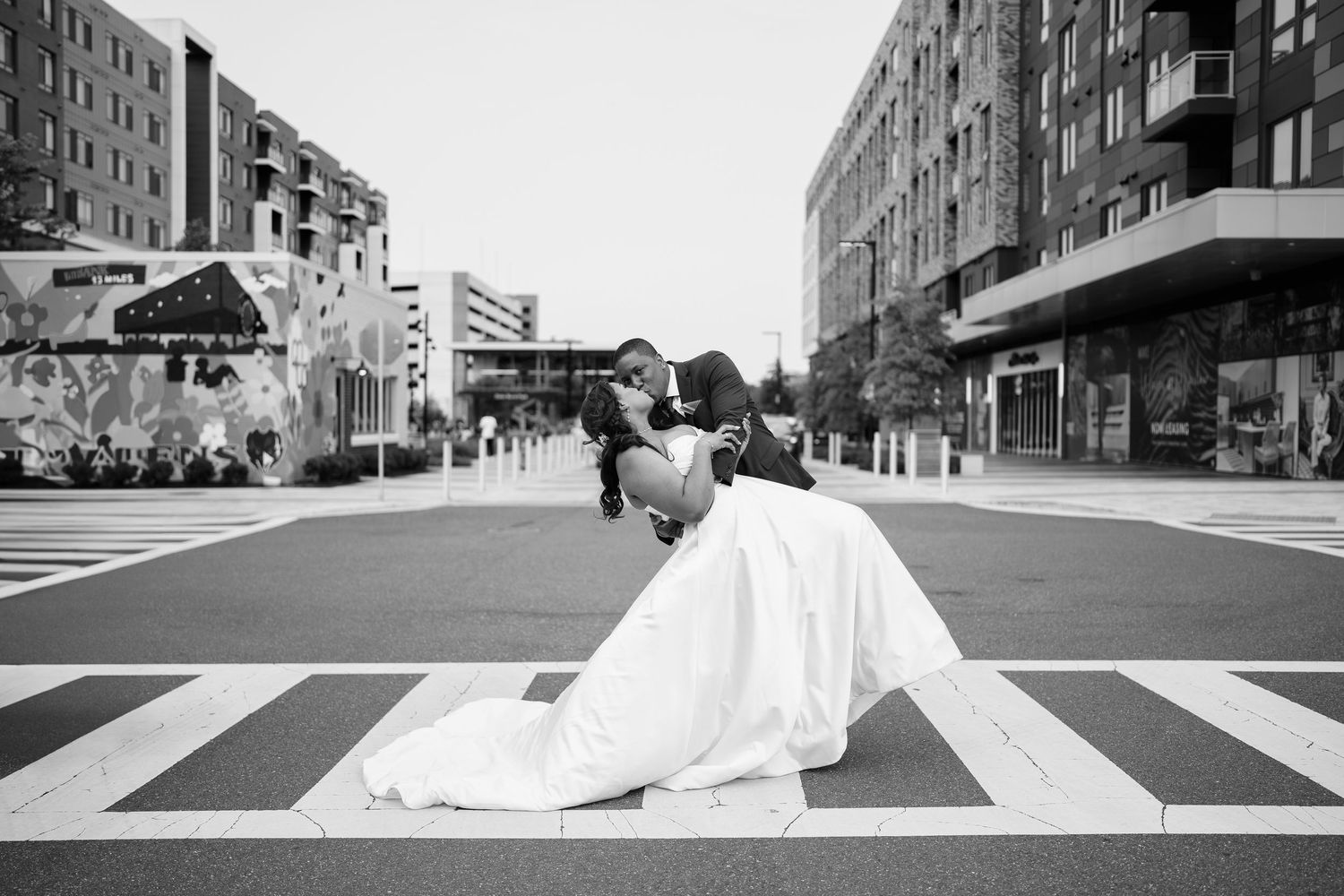 Black and white photo of a groom dipping his bride in a romantic kiss on a city crosswalk in wedding attire.