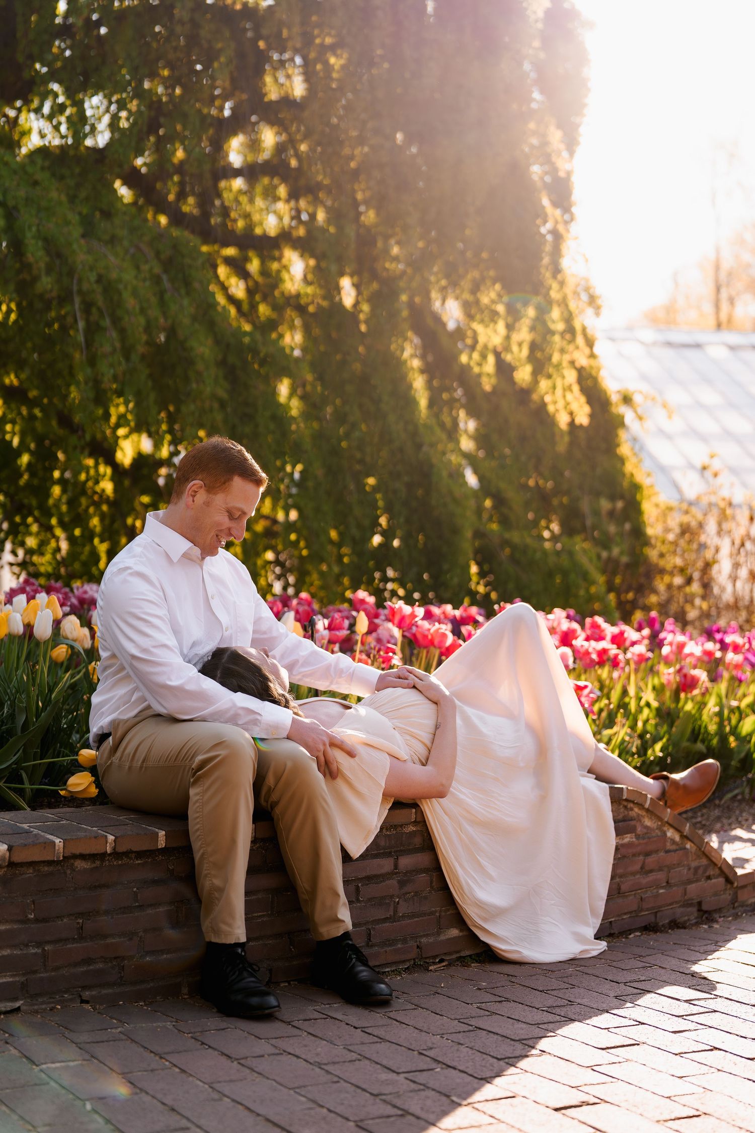 Pregnant couple sitting on brick wall surrounded by colorful tulips in sunny garden, in Brookside Gardens in Rockville, MD