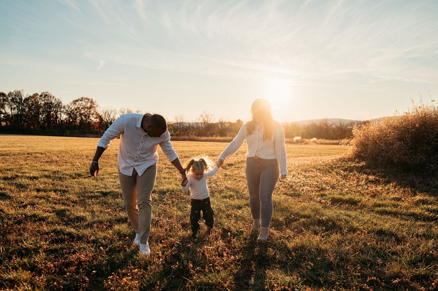 Family of three holding hands walking through a sunny golden field at sunset in autumn, in Frederick, MD