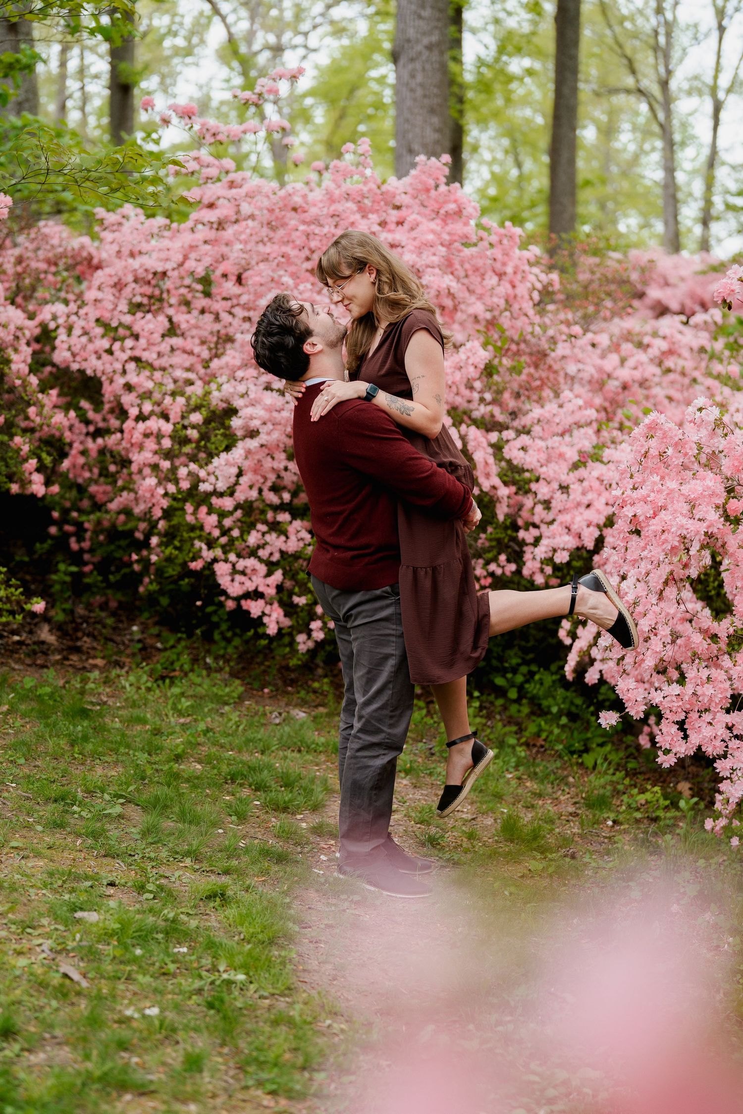 Couple sharing a romantic kiss among blooming pink azalea bushes in a lush garden during spring, during engagement session in Brookeville,MD