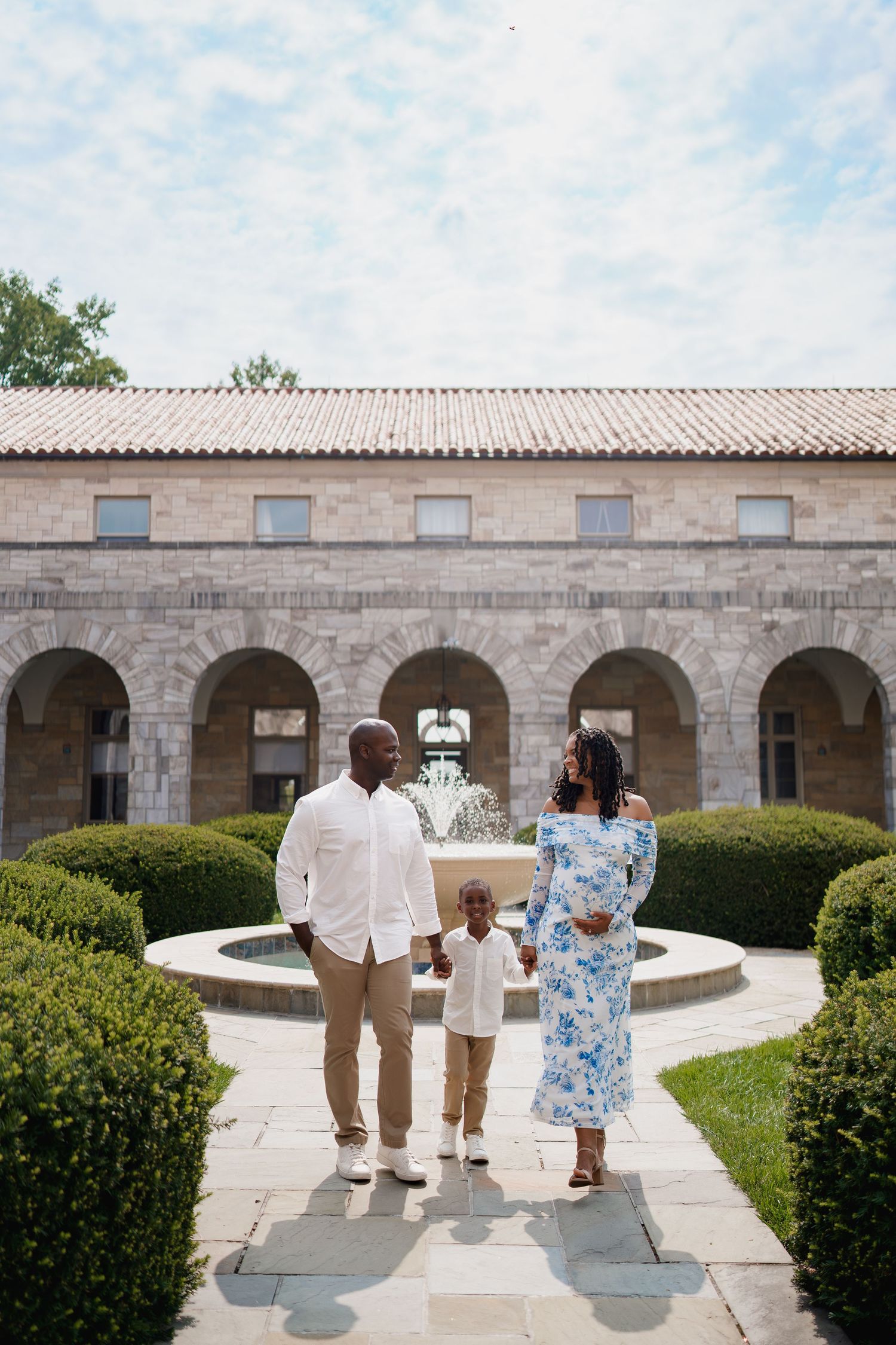 A Black family of three poses in a courtyard garden with a fountain and stone arched building backdrop, in Ellicott City, MD