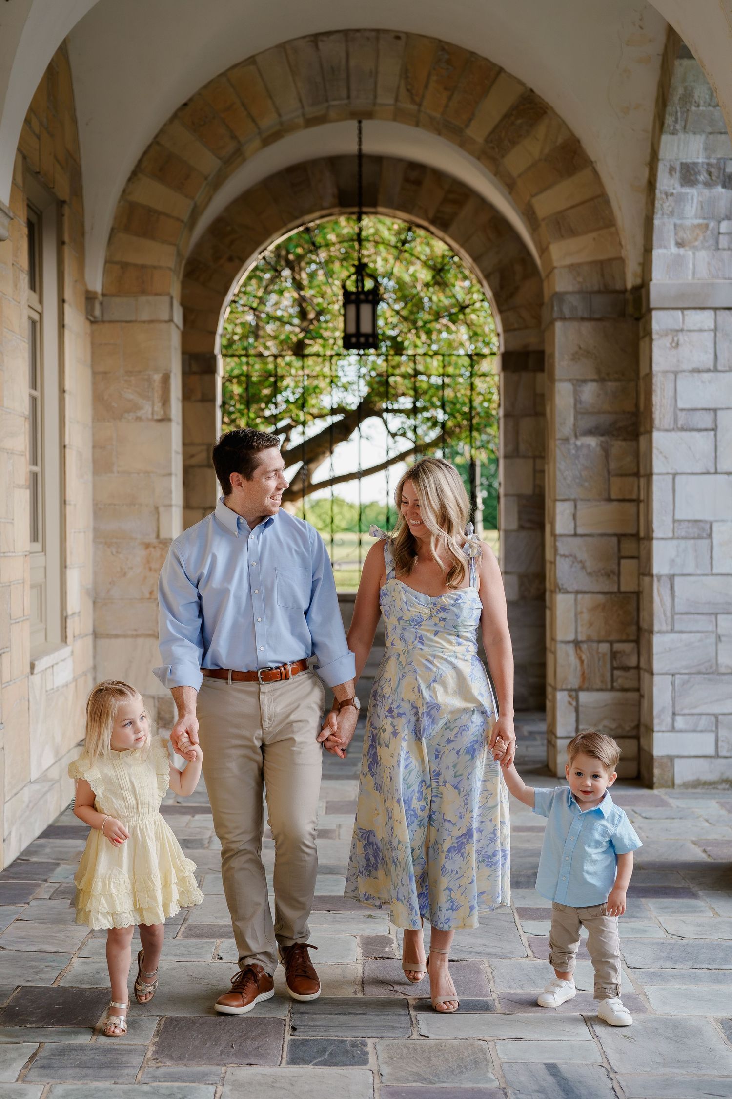 Family of four walking under stone archway, parents holding hands with young children in coordinated outfits, in Ellicott City, MD
