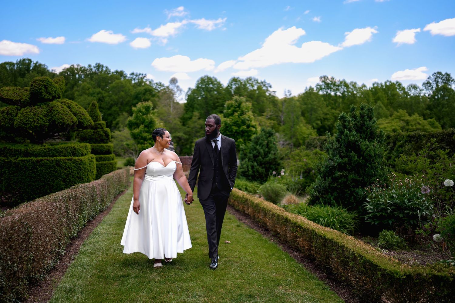 Bride and groom walk hand-in-hand through lush garden hedgerows under blue sky on their wedding day.