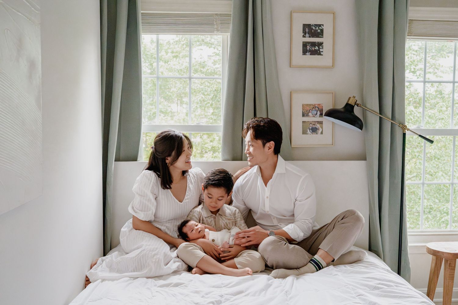 Happy Asian family with two children sitting together on a white bed near bright windows in a cozy room, in Fairfax, VA