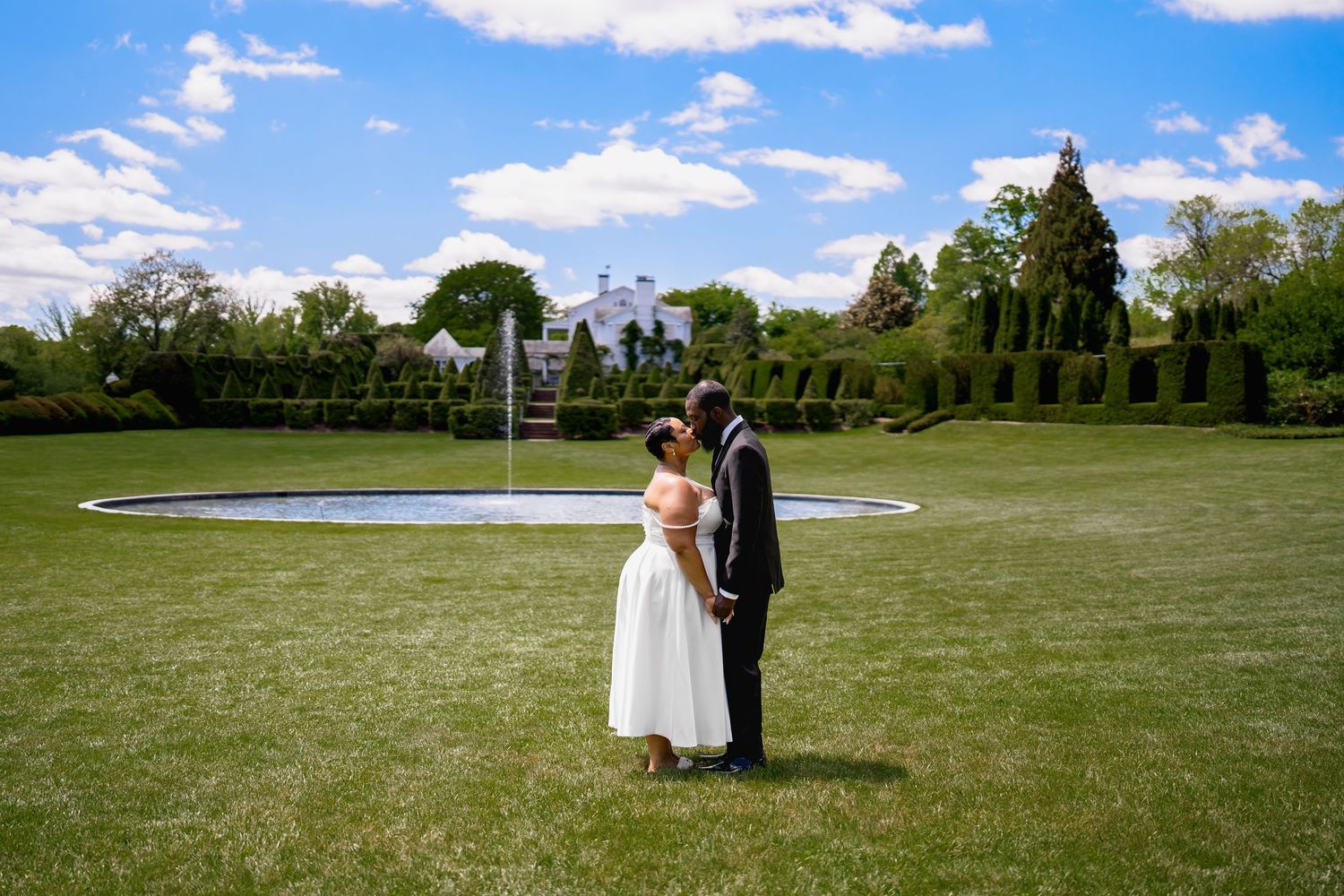 Bride and groom share a romantic moment on lush green lawn with fountain and manicured gardens on sunny day, in Baltimore, MD