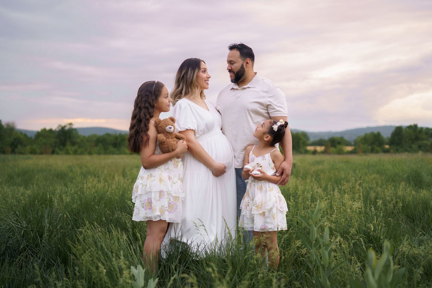 Pregnant woman in white dress with husband and two young daughters posing in green field at sunset, in Frederick, MD