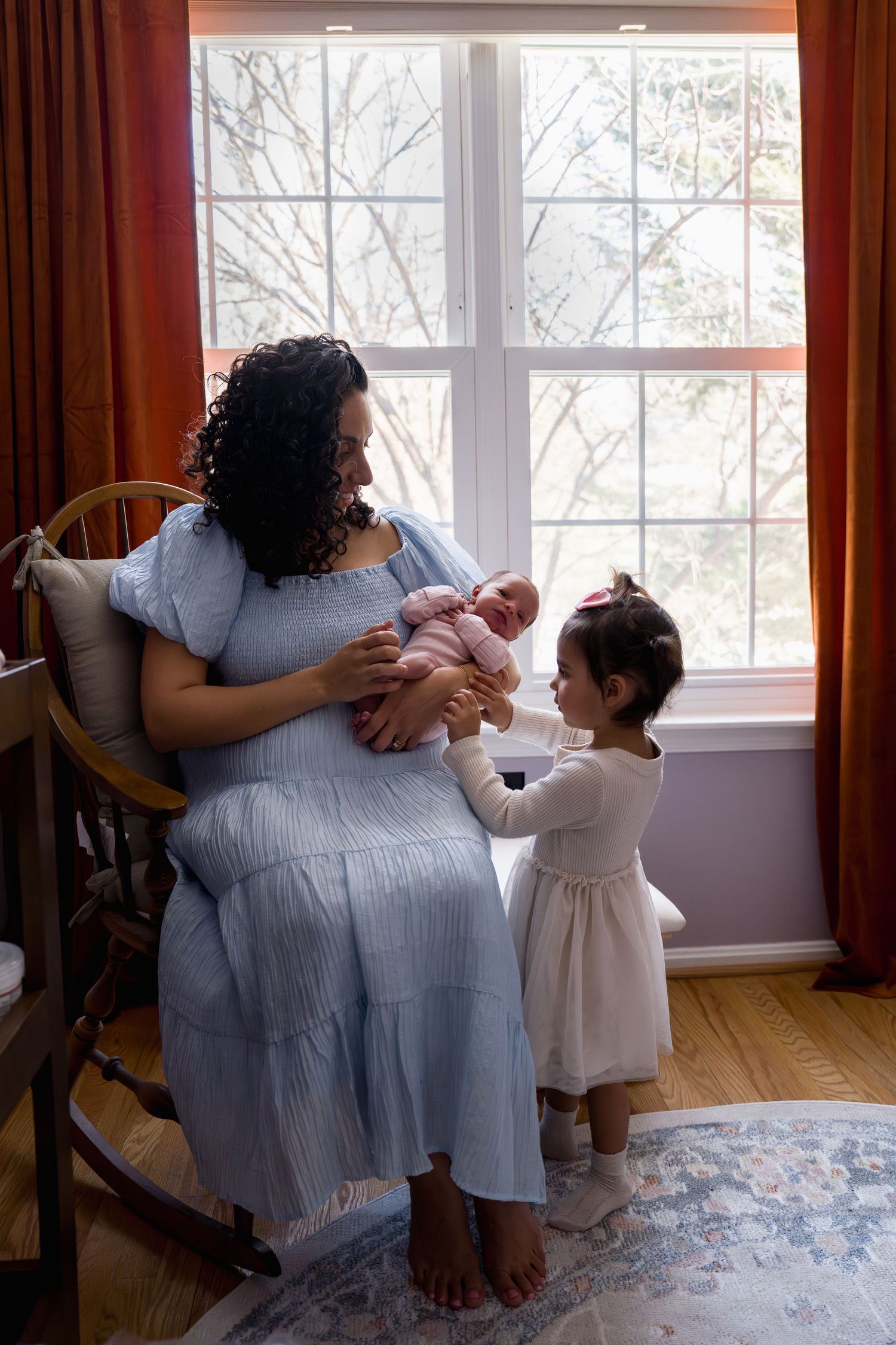 A mother in a blue dress sits in a rocking chair holding a newborn while a toddler girl reaches up to touch the baby, in Gaithersburg, MD
