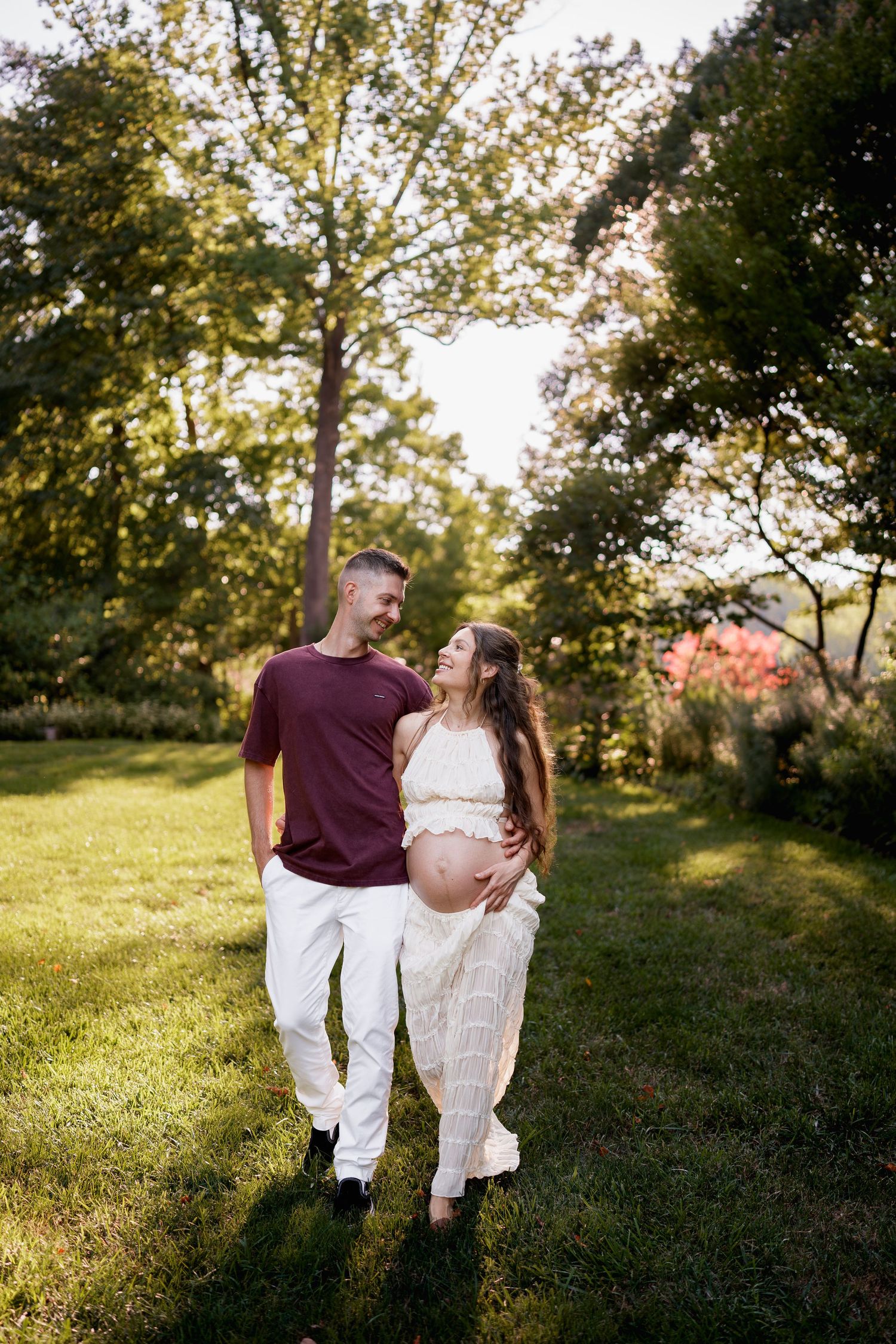 Pregnant couple walking in lush green garden, woman in white dress cradling baby bump, man in burgundy shirt smiling, in Wheaton, MD