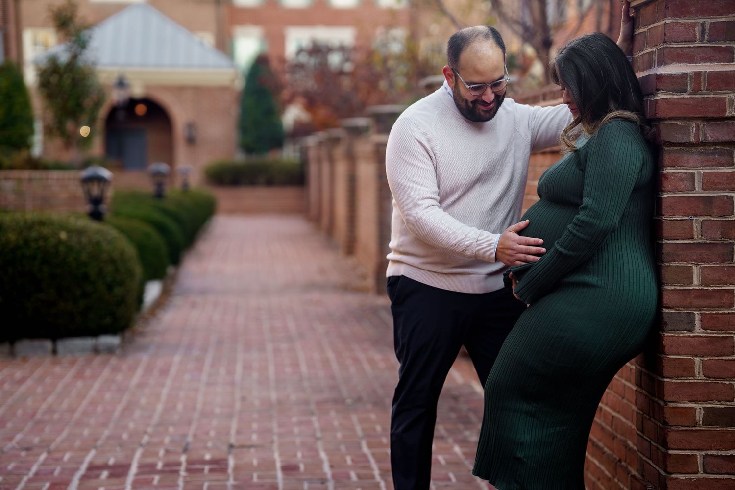 A man tenderly touches a pregnant woman's belly as they share a loving moment against a brick wall outdoors, in Alexandria, VA