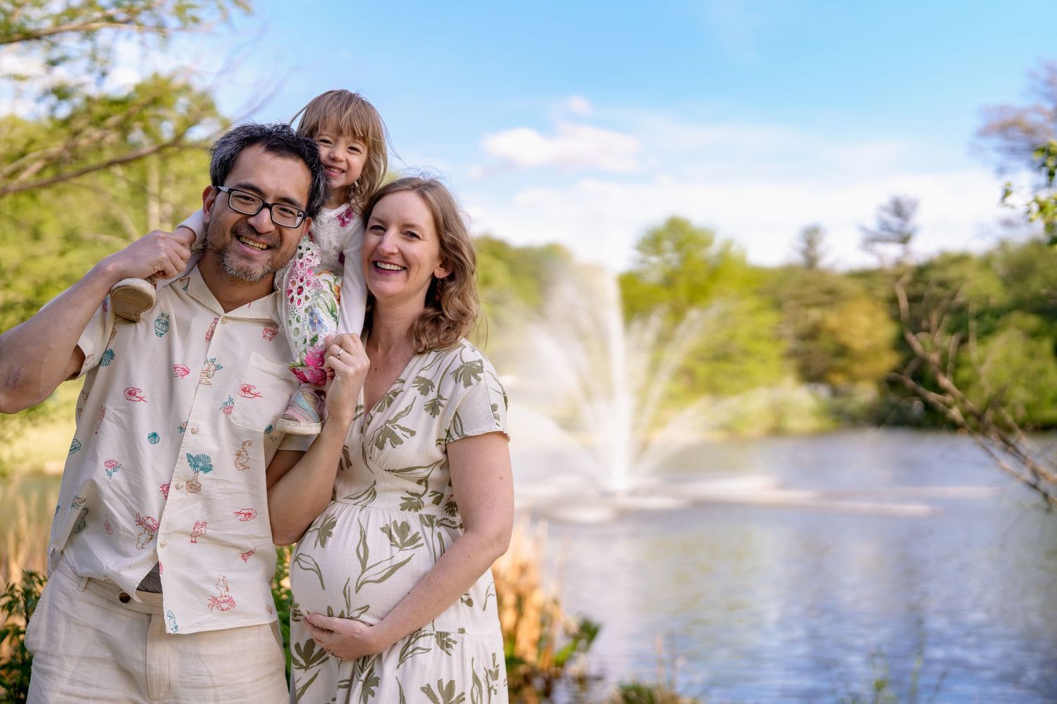 Smiling pregnant woman with husband and young daughter posing by a serene fountain outdoors on a sunny day, in Northern Virginia