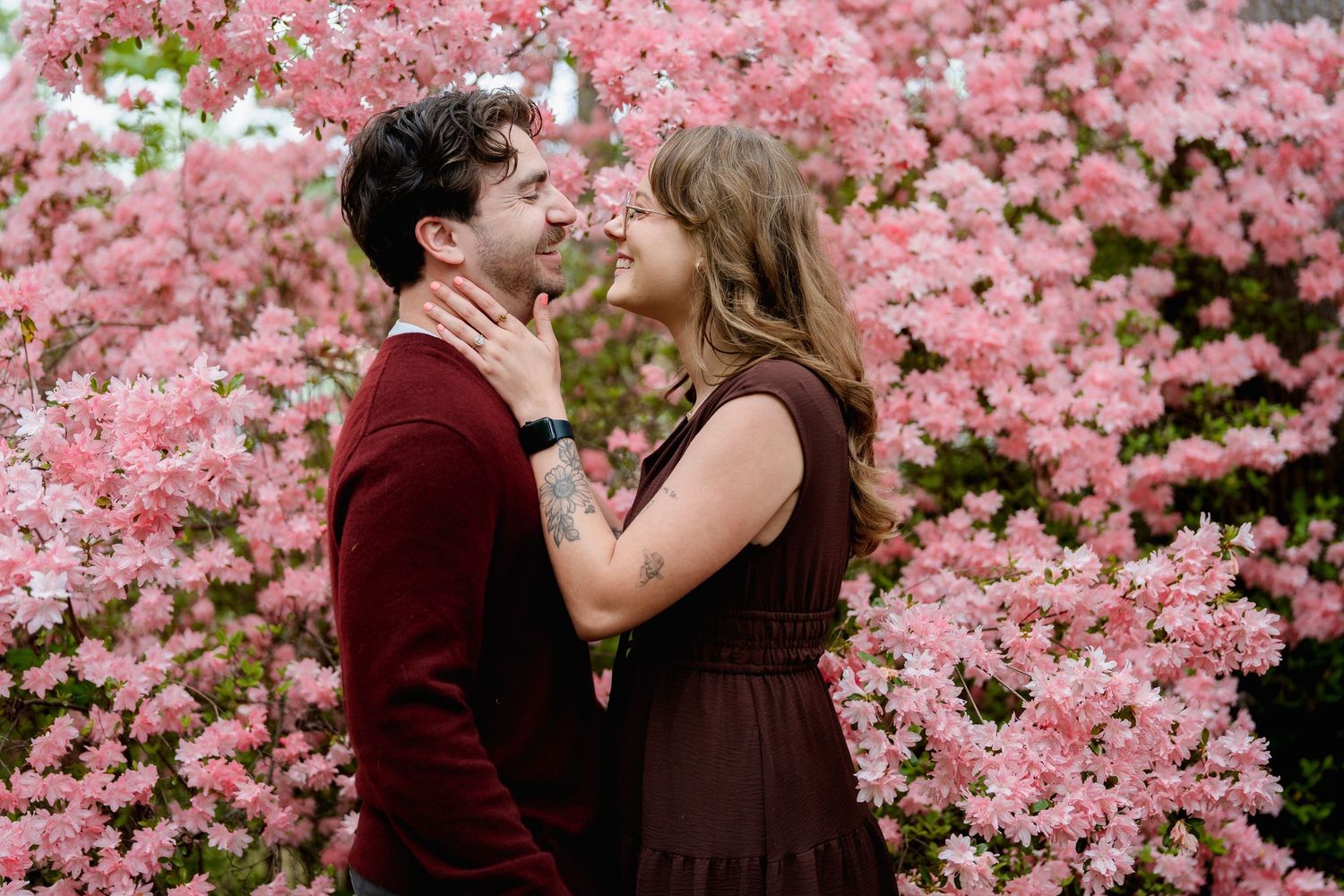 Couple sharing a romantic moment in front of blooming pink azalea bushes, wearing matching burgundy outfits, in Brookeville, MD