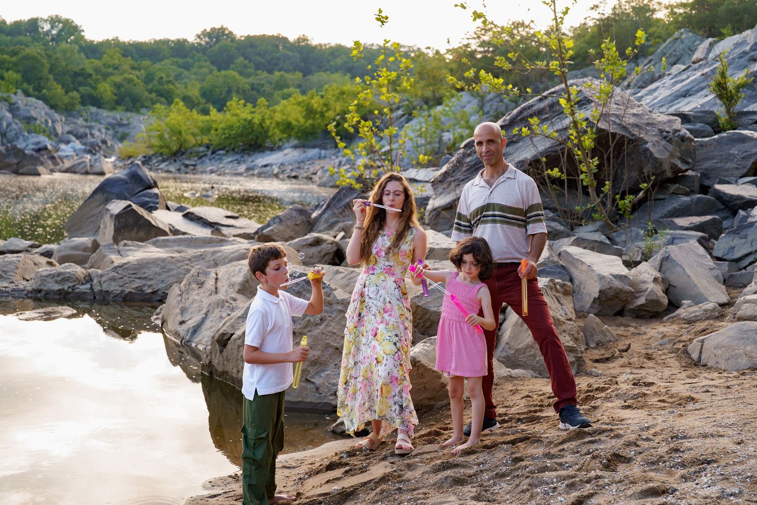 Family of five posing on rocky riverbank surrounded by lush green trees during golden hour sunset, in Great Falls Park, in Maryland
