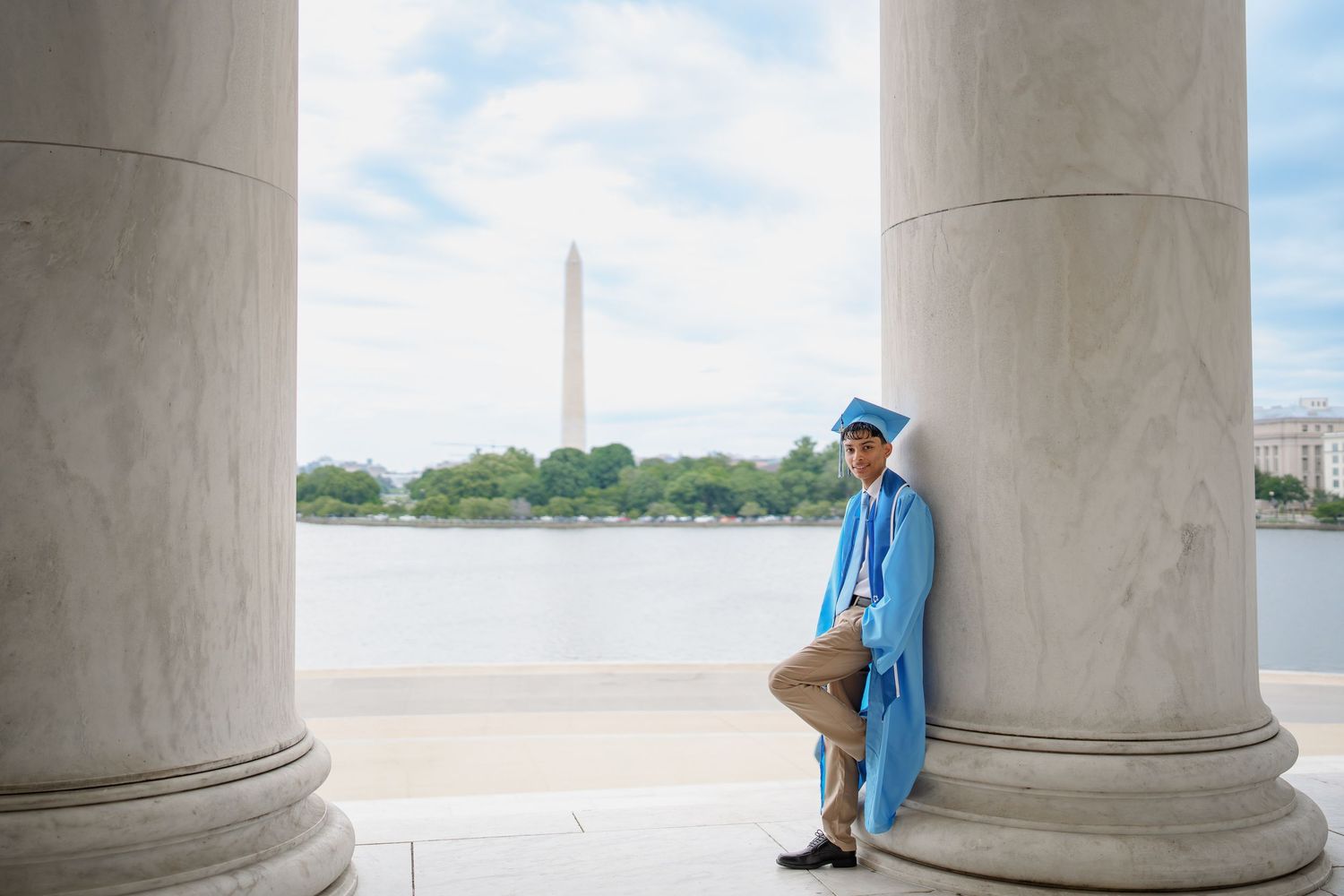 Graduate in blue cap and gown poses between marble columns at Jefferson Memorial with Washington Monument in background, in DC