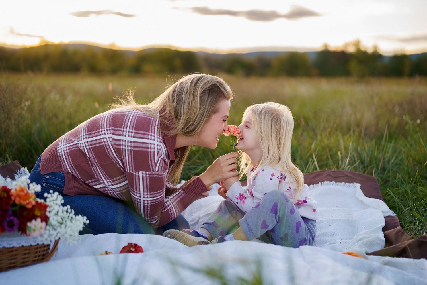 A mother and daughter share a tender moment smelling a flower during a golden hour picnic in a meadow, a family session in Frederick, MD