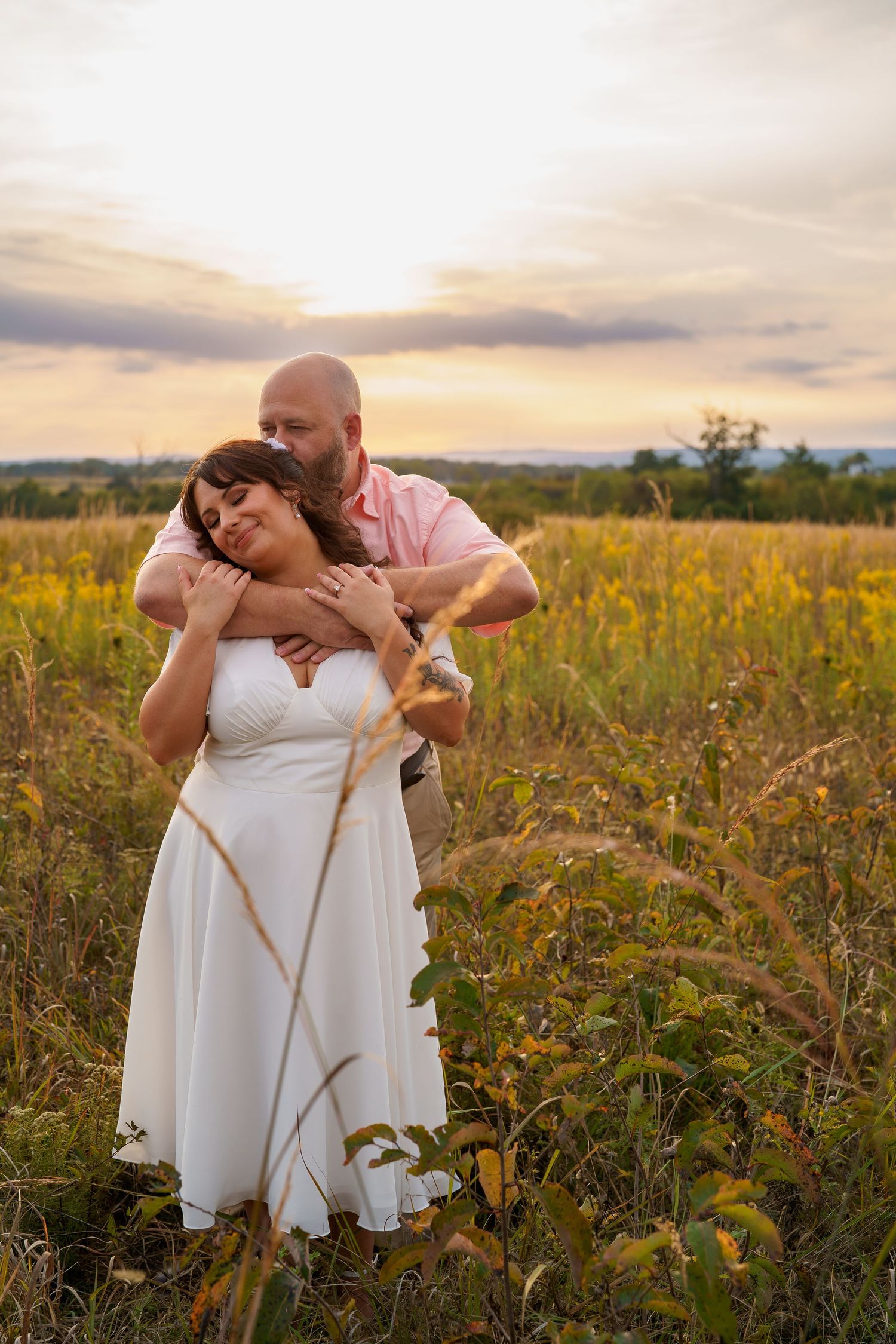 Couple embracing in golden field at sunset, bride in white dress, groom in pink shirt, romantic outdoor wedding photo, in Gettysburg, PA