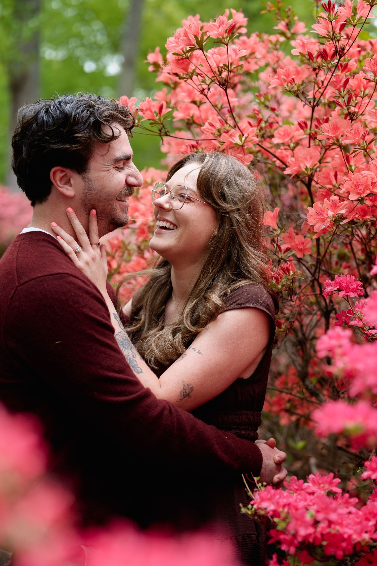 Couple embracing and laughing among vibrant pink azalea blooms in a lush garden setting, engagement session in Brookeville, MD