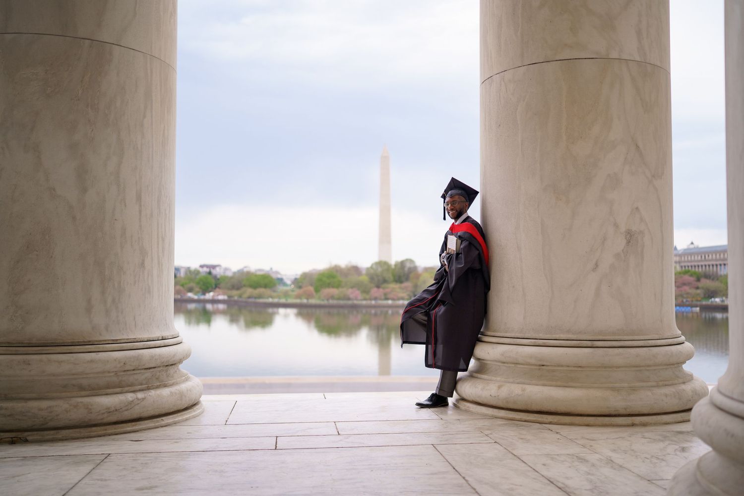 Graduate in cap and gown stands between columns at the Jefferson Memorial with Washington Monument in background in washington DC