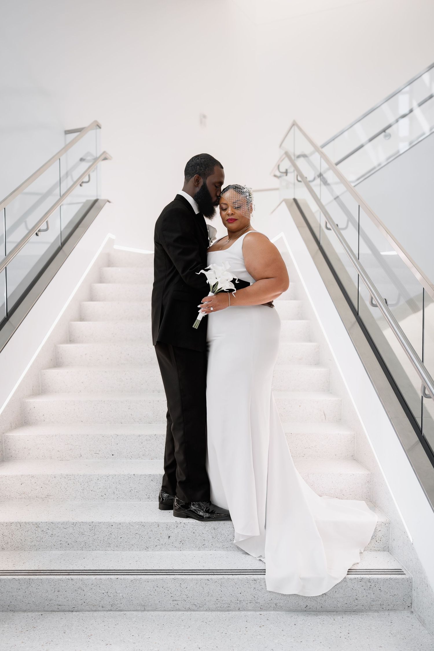 Elegant Black couple in wedding attire embrace on white marble stairs, bride holding white bouquet in sleek gown.