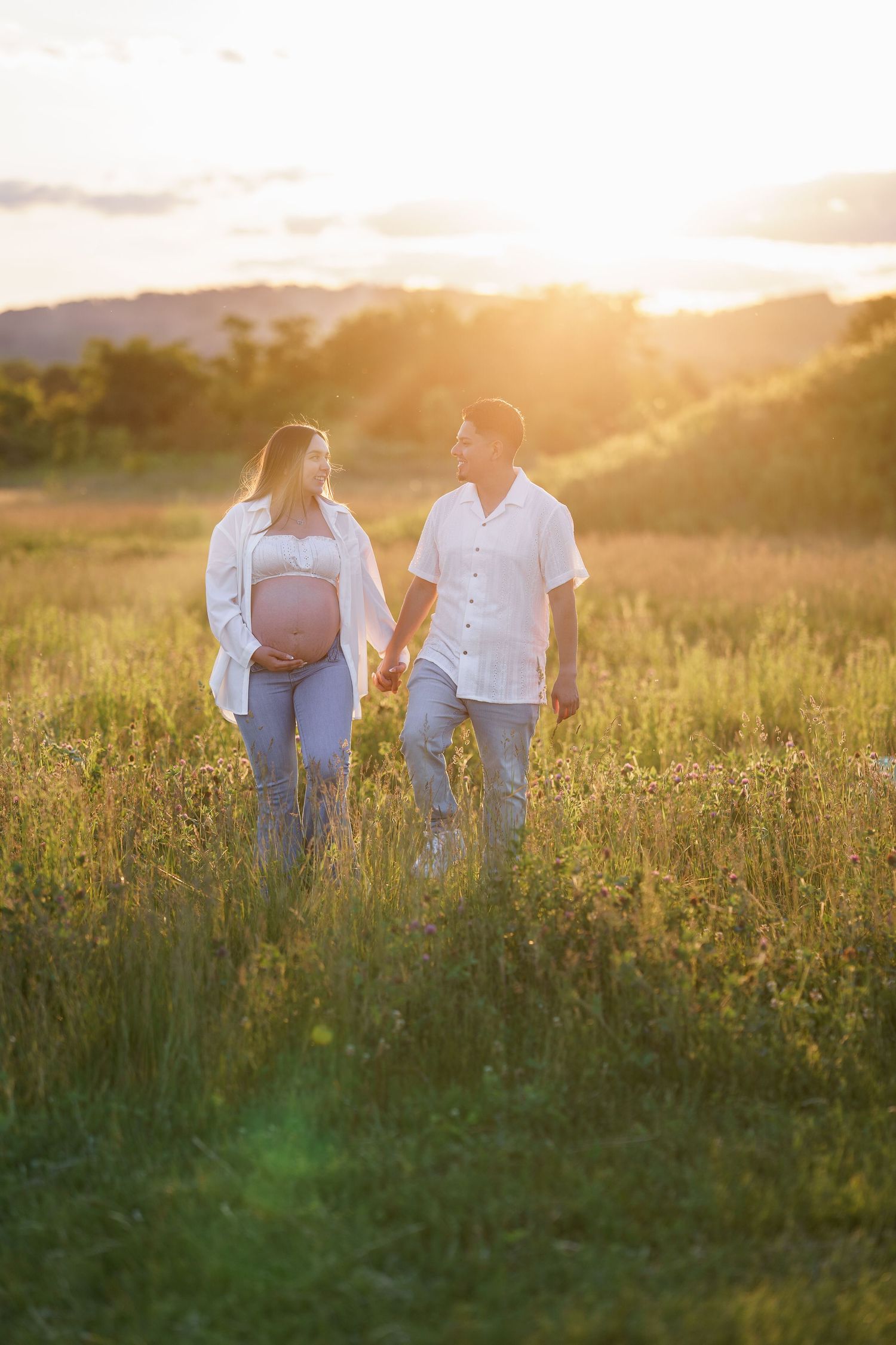Pregnant couple holding hands in golden sunset field, woman cradling baby bump, both smiling at each other, in Frederick, MD