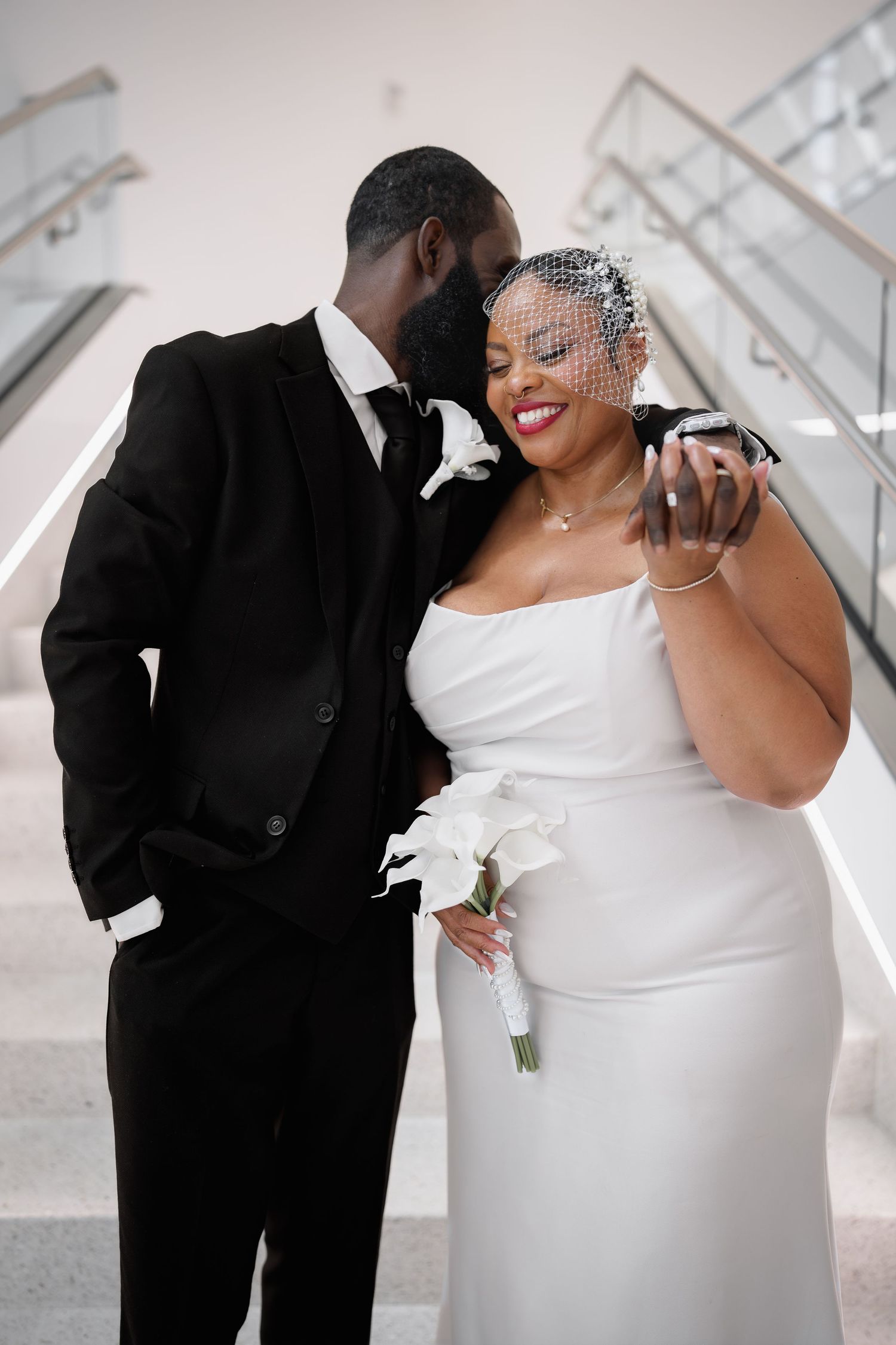 A joyful bride in a white gown and veil holds white calla lilies as her groom in a black suit leans in close on stairs, in Columbia, MD