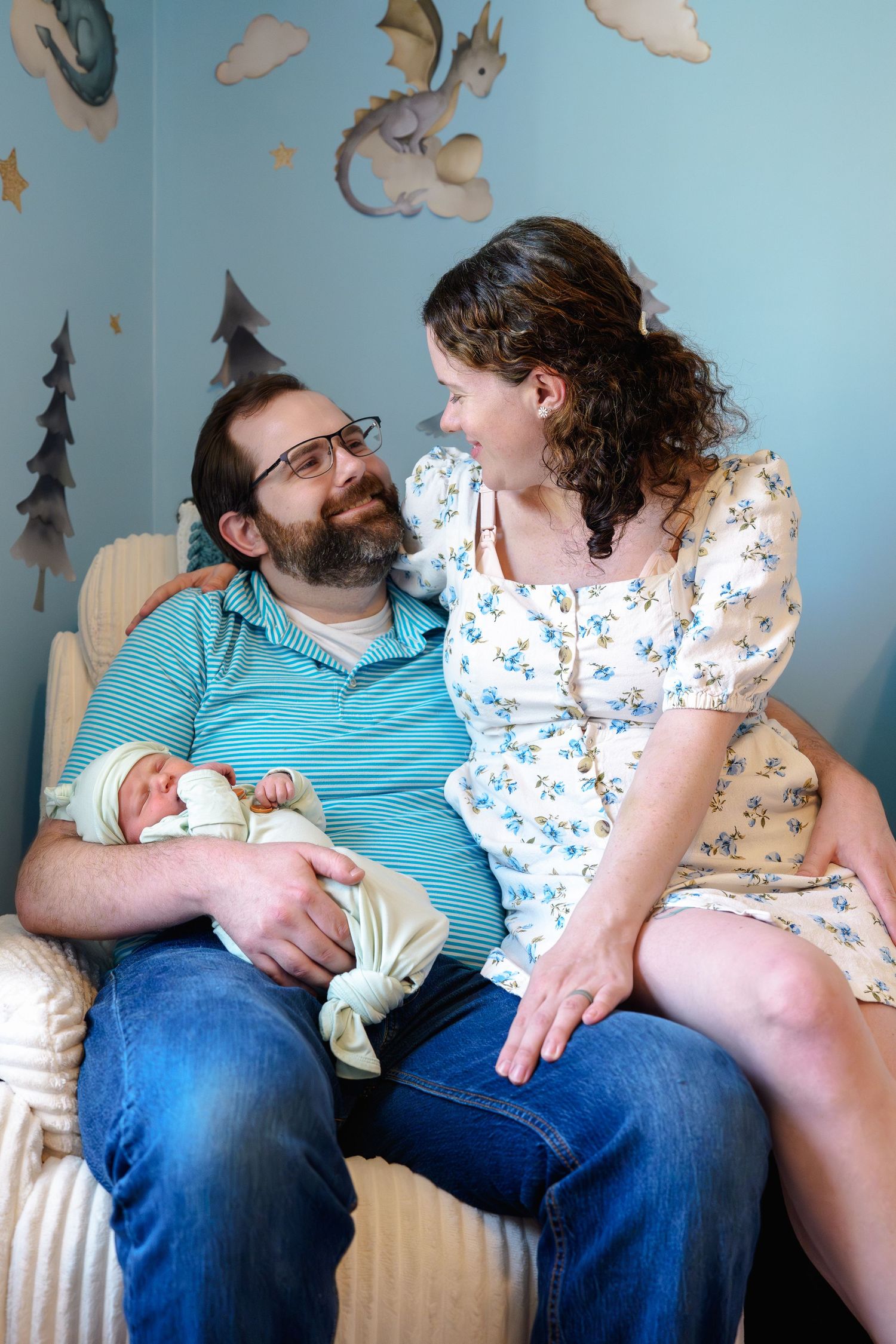 A couple smiles at each other in a nursery, father holding newborn baby in striped shirt, mother in floral dress beside him, in Maryland