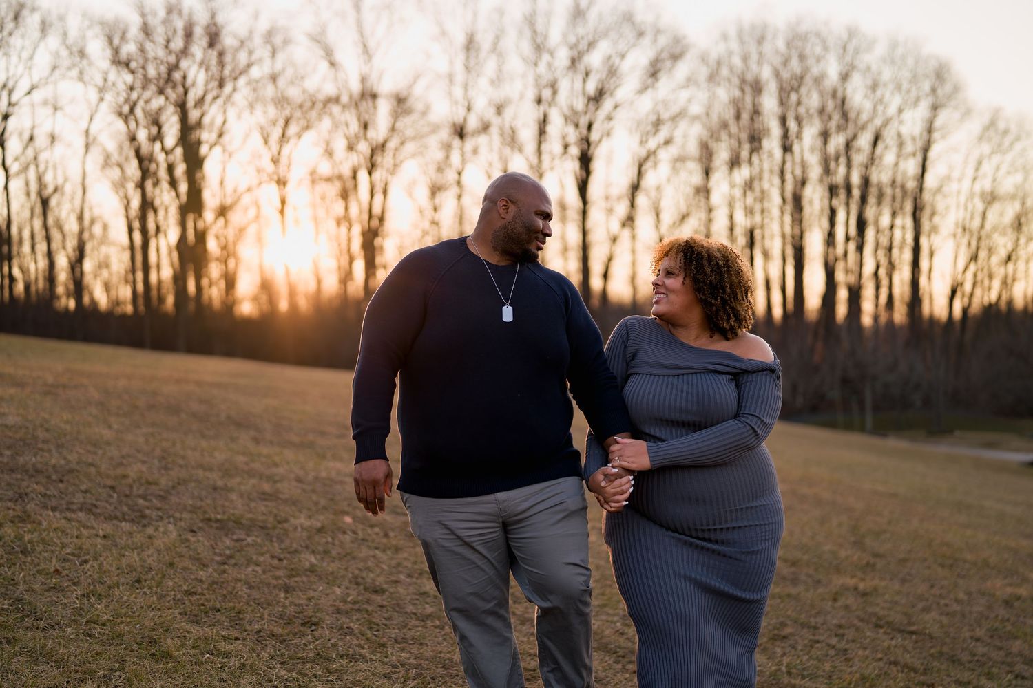 Couple walking hand in hand at sunset in a field with bare trees, sharing a loving glance, during a maternity session in Ellicott City, MD