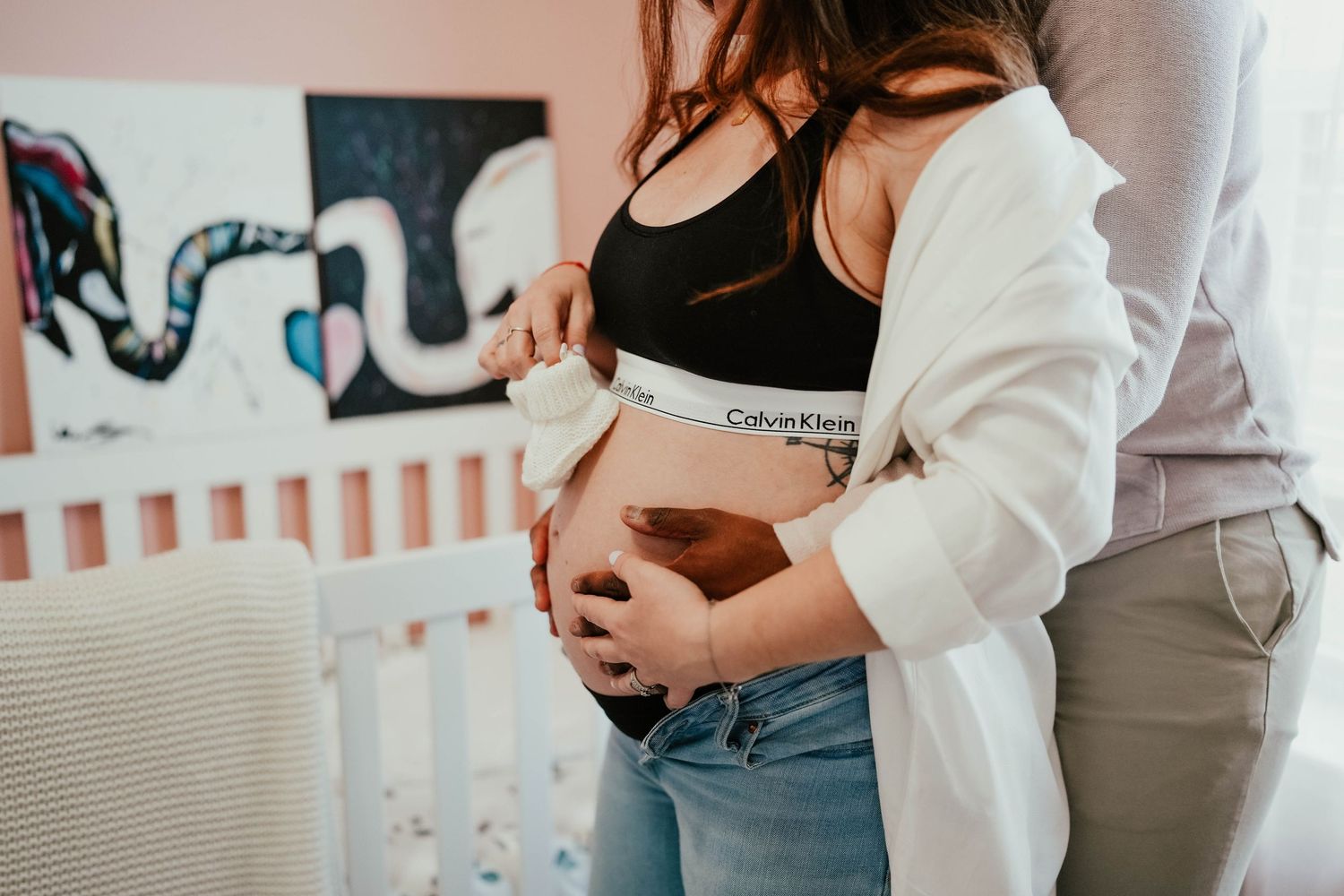 Pregnant woman in Calvin Klein bralette and jeans with partner holding baby shoes near nursery crib.