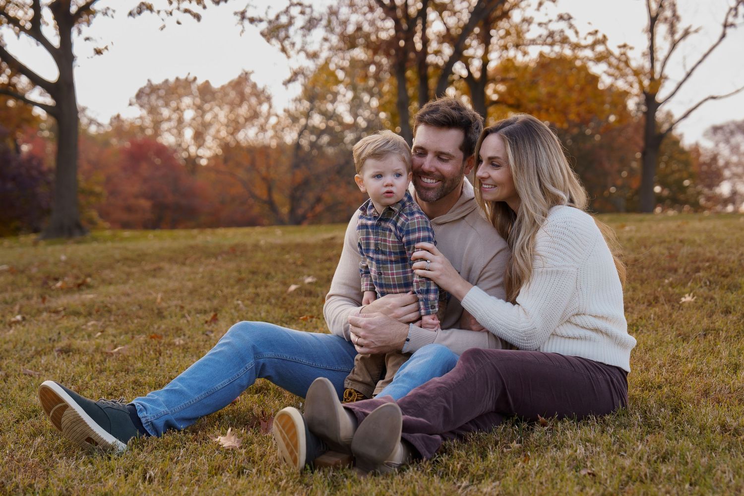 Happy family of three sitting on grass in autumn park, parents smiling at toddler son in plaid shirt, in Constitution Gardens in DC