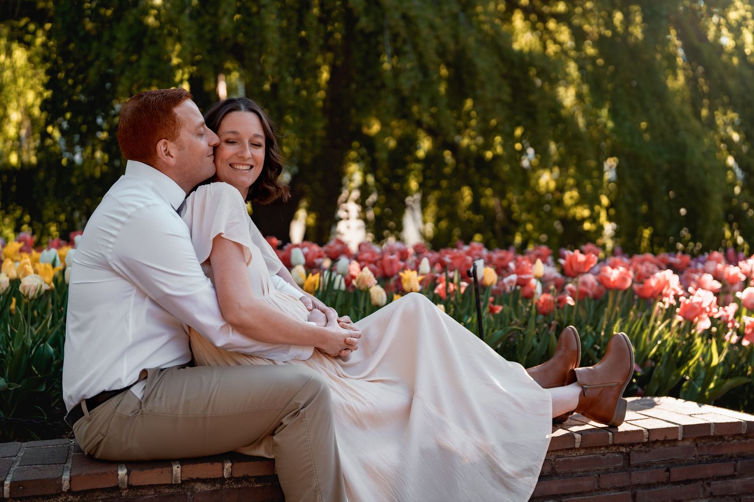 Pregnant couple sitting on brick wall surrounded by colorful tulips in a sunlit garden, in Brookside Gardens, MD during maternity session,