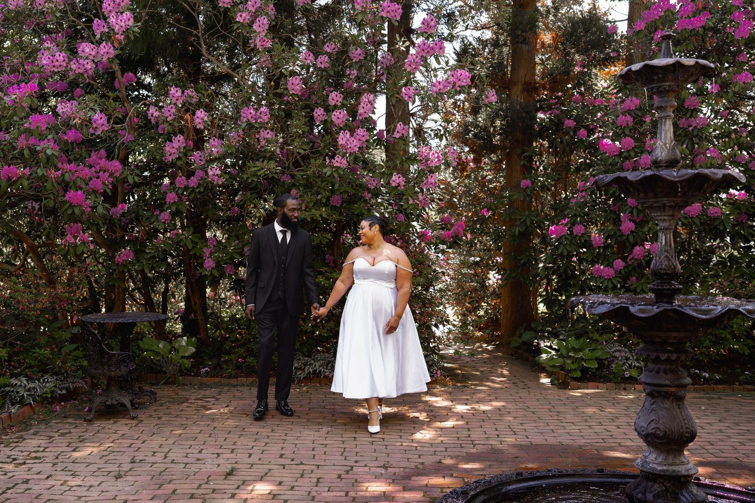 Bride and groom hold hands in a garden courtyard surrounded by blooming pink flowers and a stone fountain.