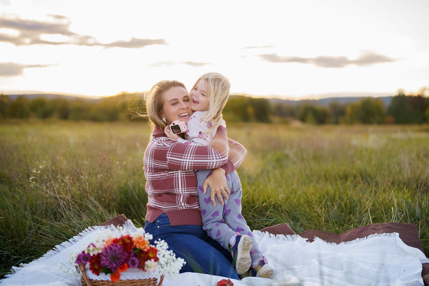A mother and young daughter share a warm embrace on a white blanket in a sunlit meadow during golden hour, in Frederick, MD