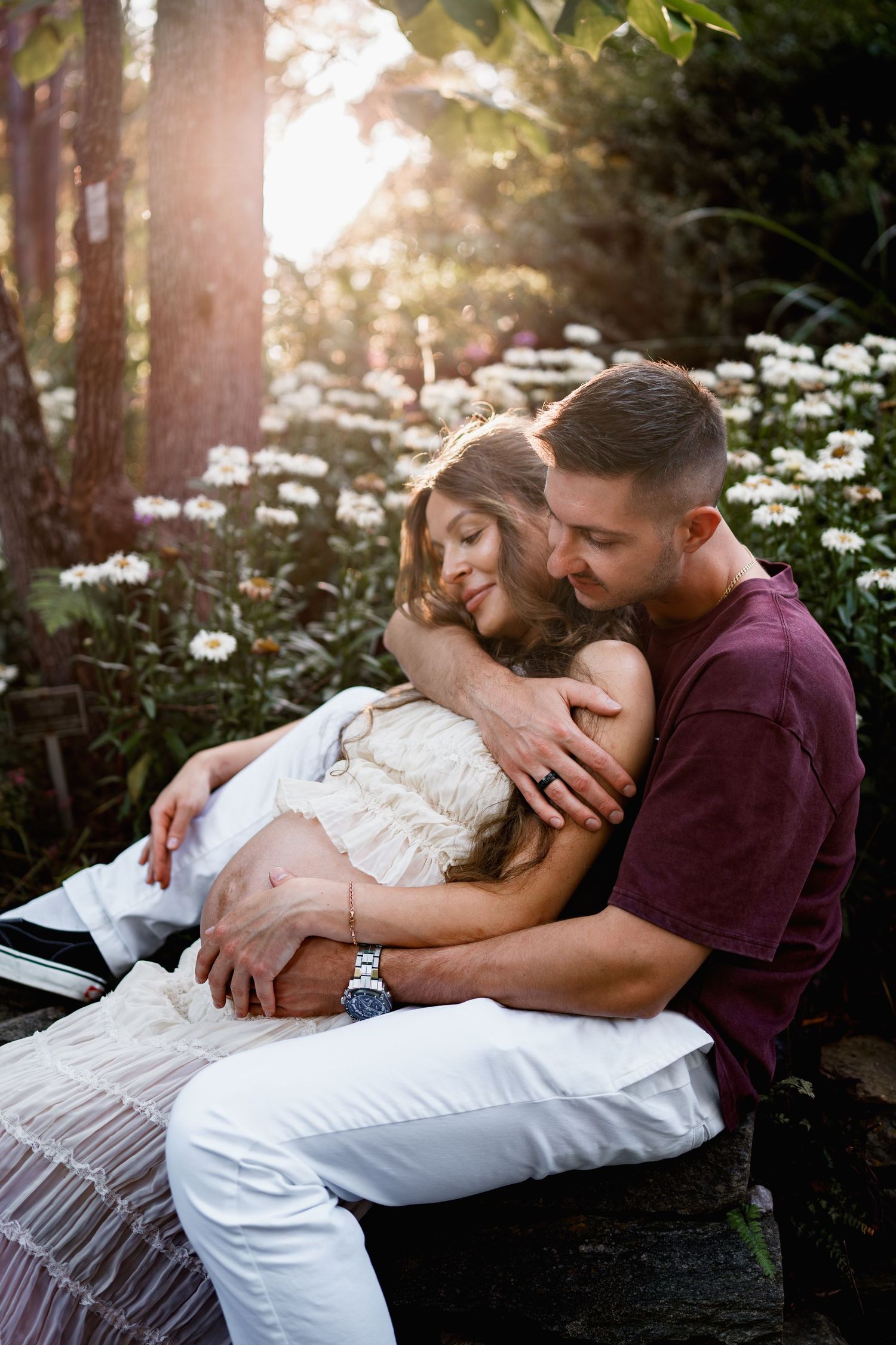 A couple shares a tender embrace on a garden bench surrounded by white daisies and warm sunlight filtering through trees, in Maryland