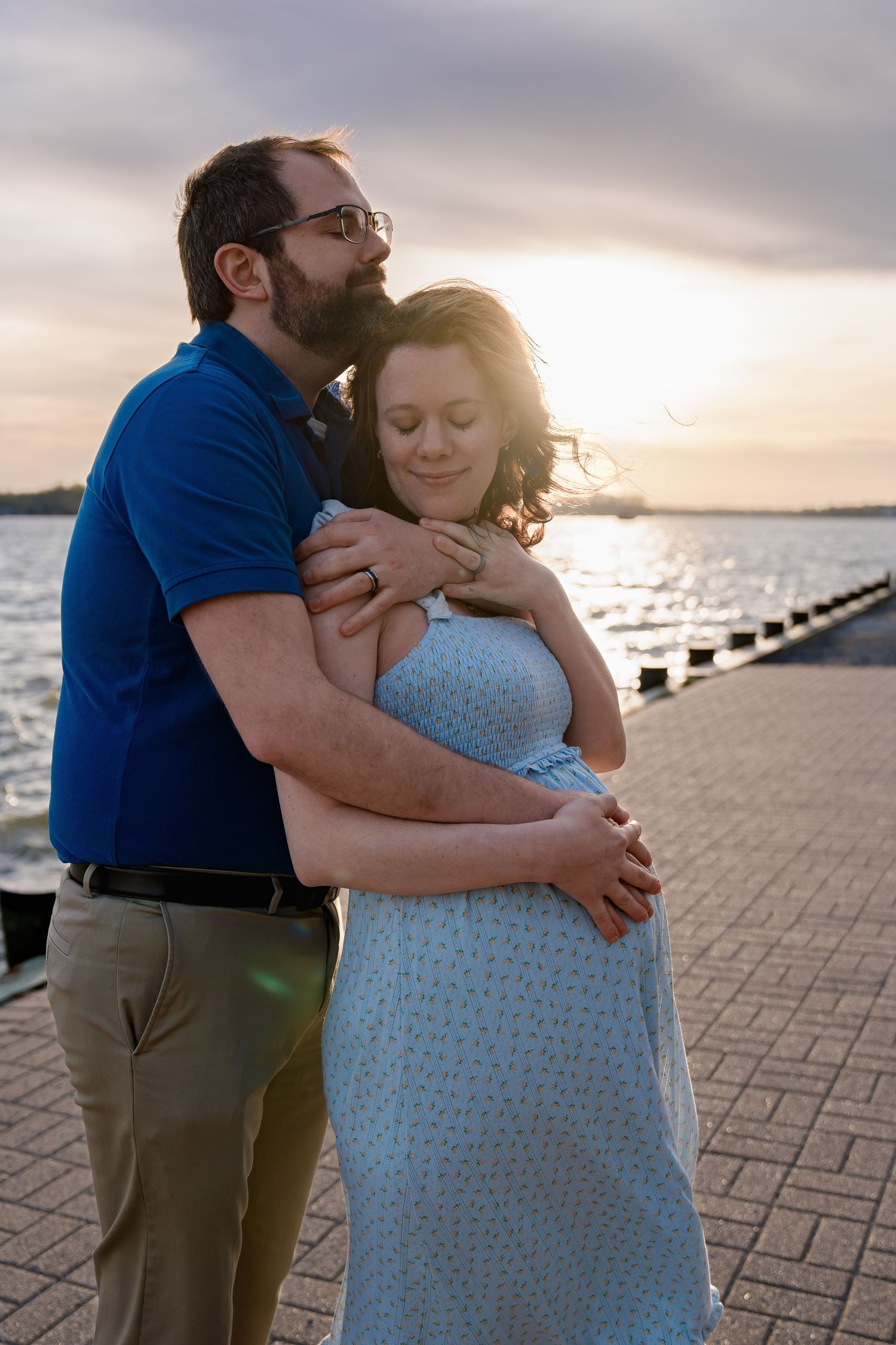 A couple embraces at sunset by a waterfront pier, the man hugging a pregnant woman in a blue dress from behind in Annapolis, Maryland
