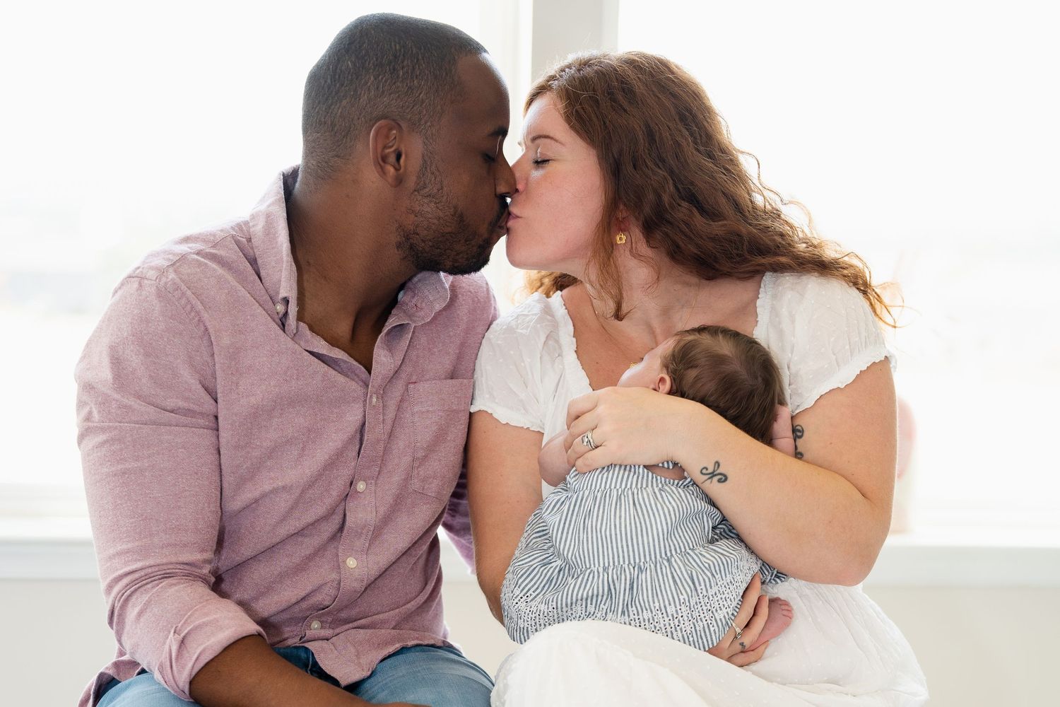 A loving couple shares a tender kiss while holding their sleeping baby in a bright, peaceful home setting, in Frederick MD