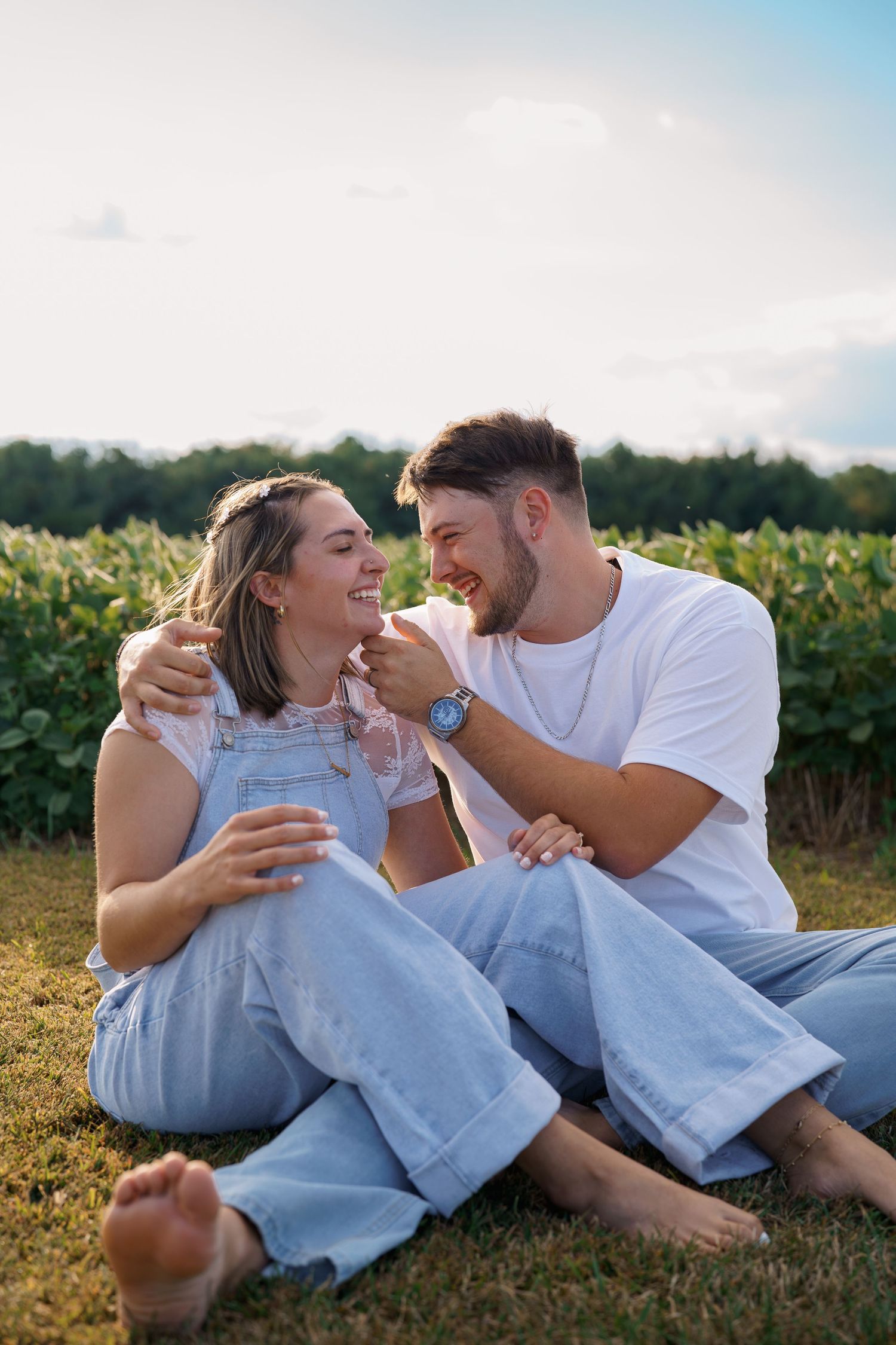 Happy couple sitting barefoot on grass near green fields, laughing and embracing on a sunny day.