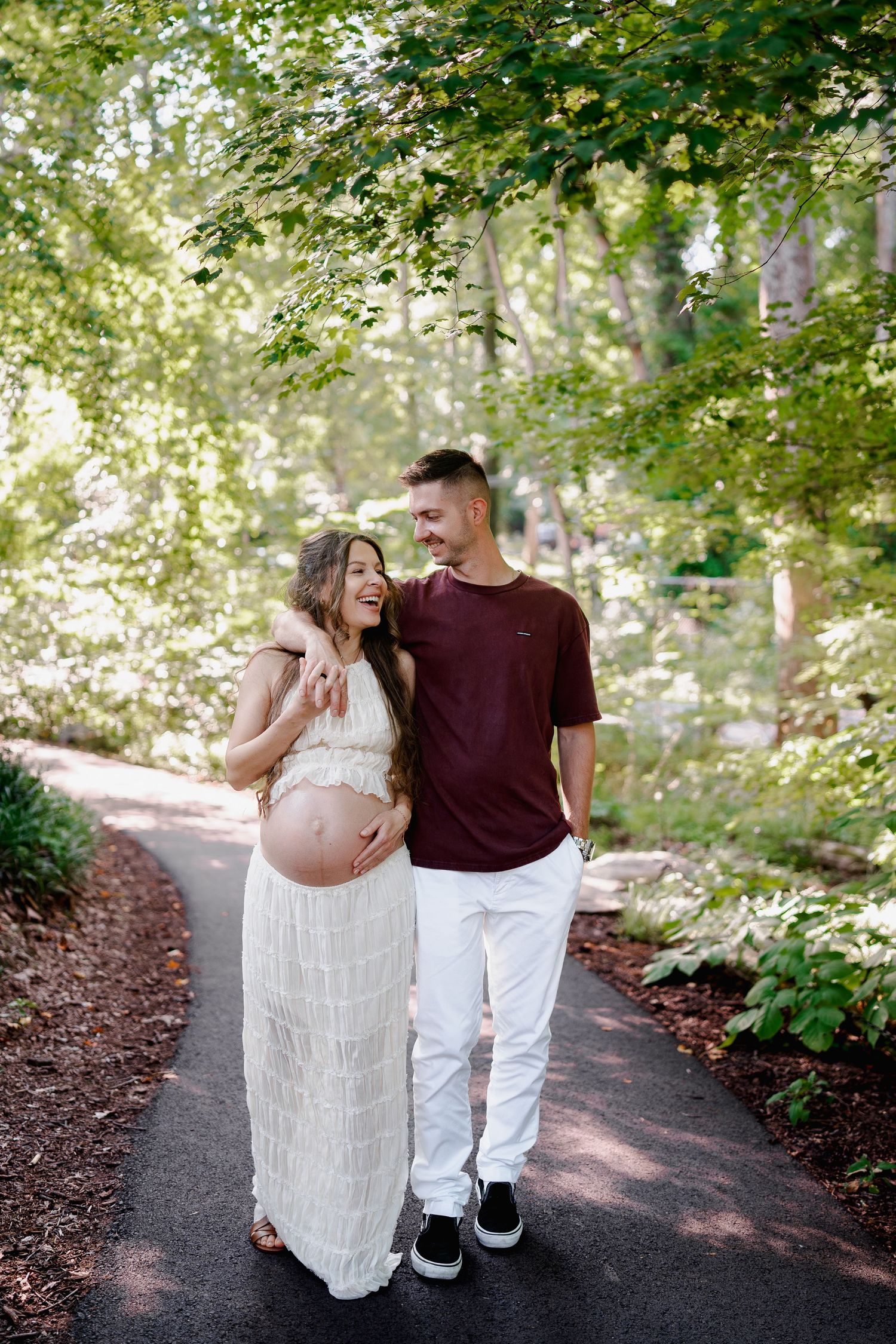Smiling pregnant couple walking on a tree-lined path, woman in white dress holding baby bump, man in maroon shirt and white pants, in MD