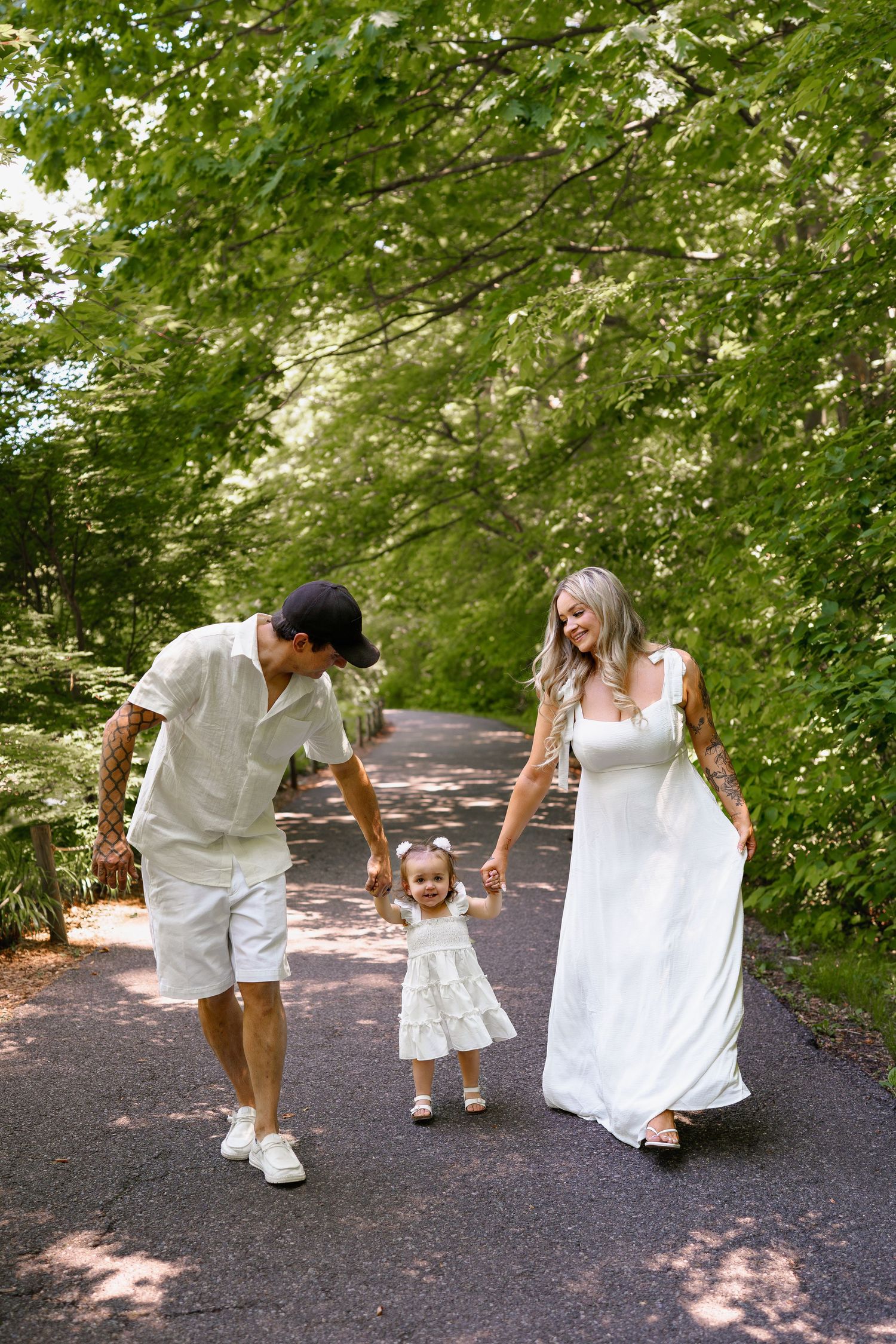A happy family walks hand-in-hand on a tree-lined path, parents guiding their toddler daughter in matching white outfits, in Maryland