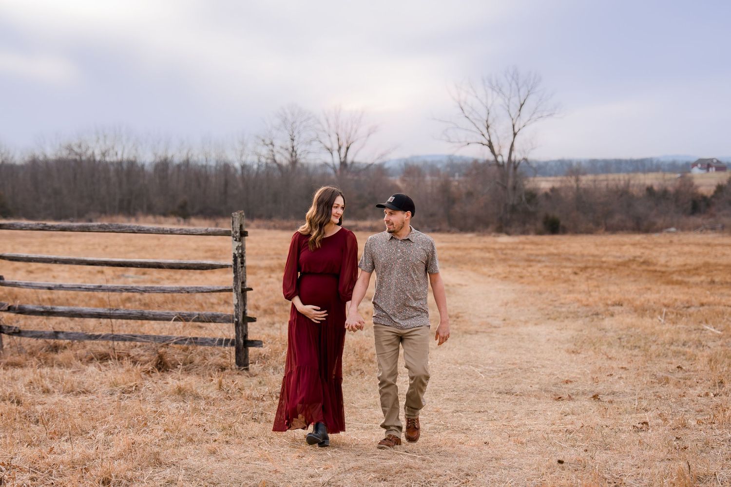 Pregnant couple walks hand-in-hand on a rural path, woman in red maternity gown, man in casual attire, in Gettysburg, PA