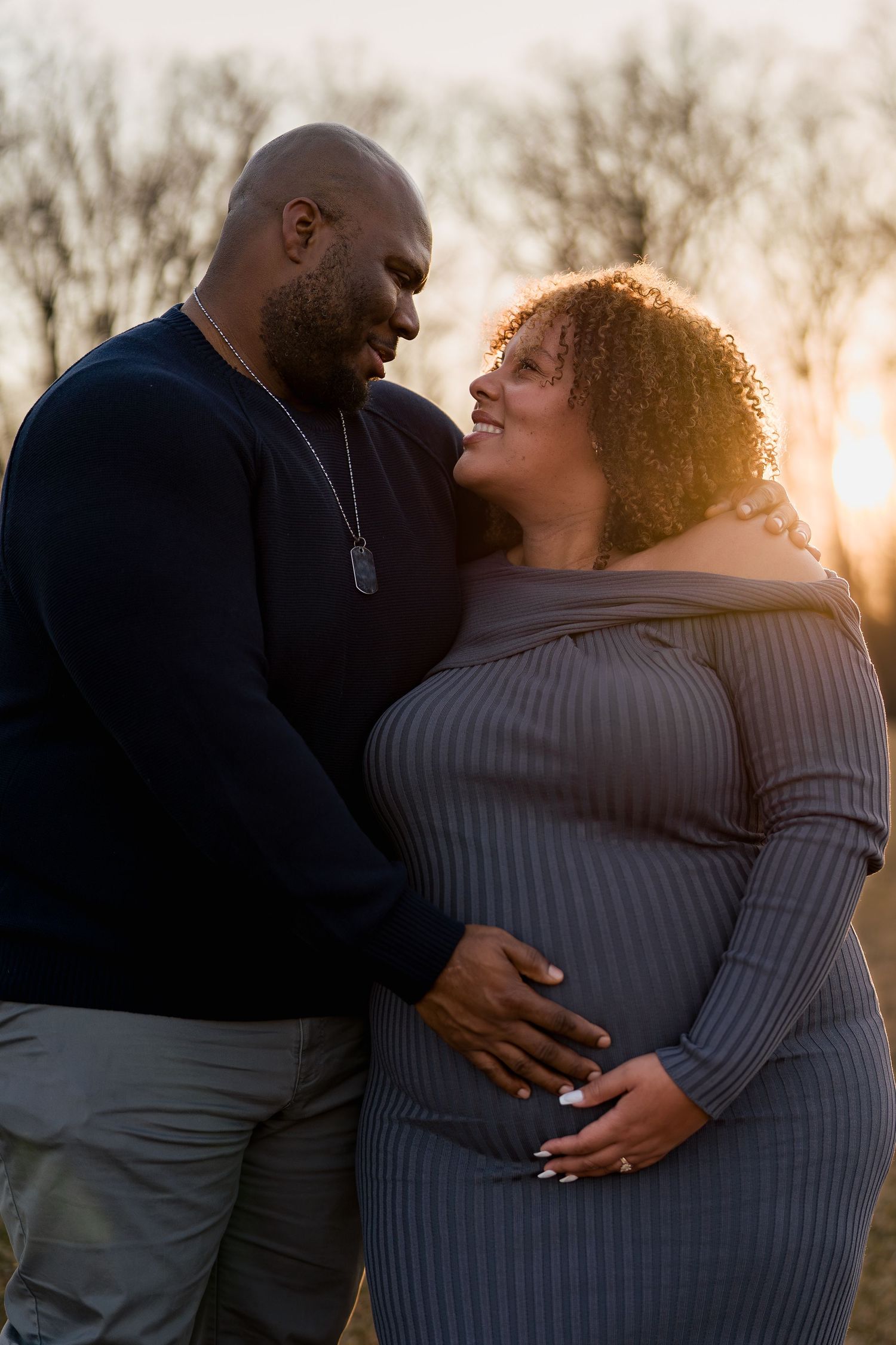 A couple shares a tender moment at sunset, with the woman in a gray dress cradling her pregnant belly outdoors, in Ellicott City, MD