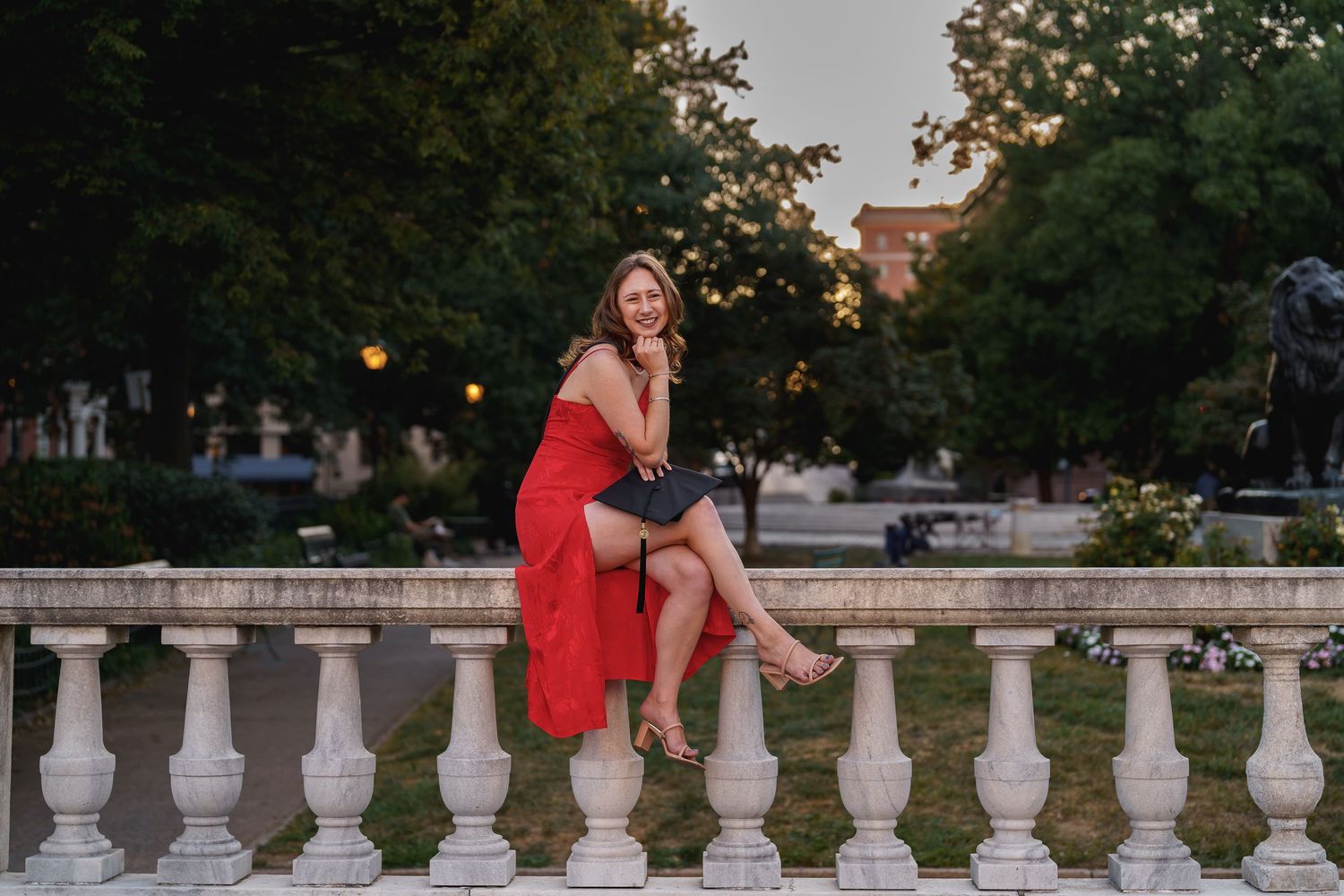Woman in red dress sitting on white balustrade in park at dusk, smiling elegantly during a graduation session in Baltimore, Maryland