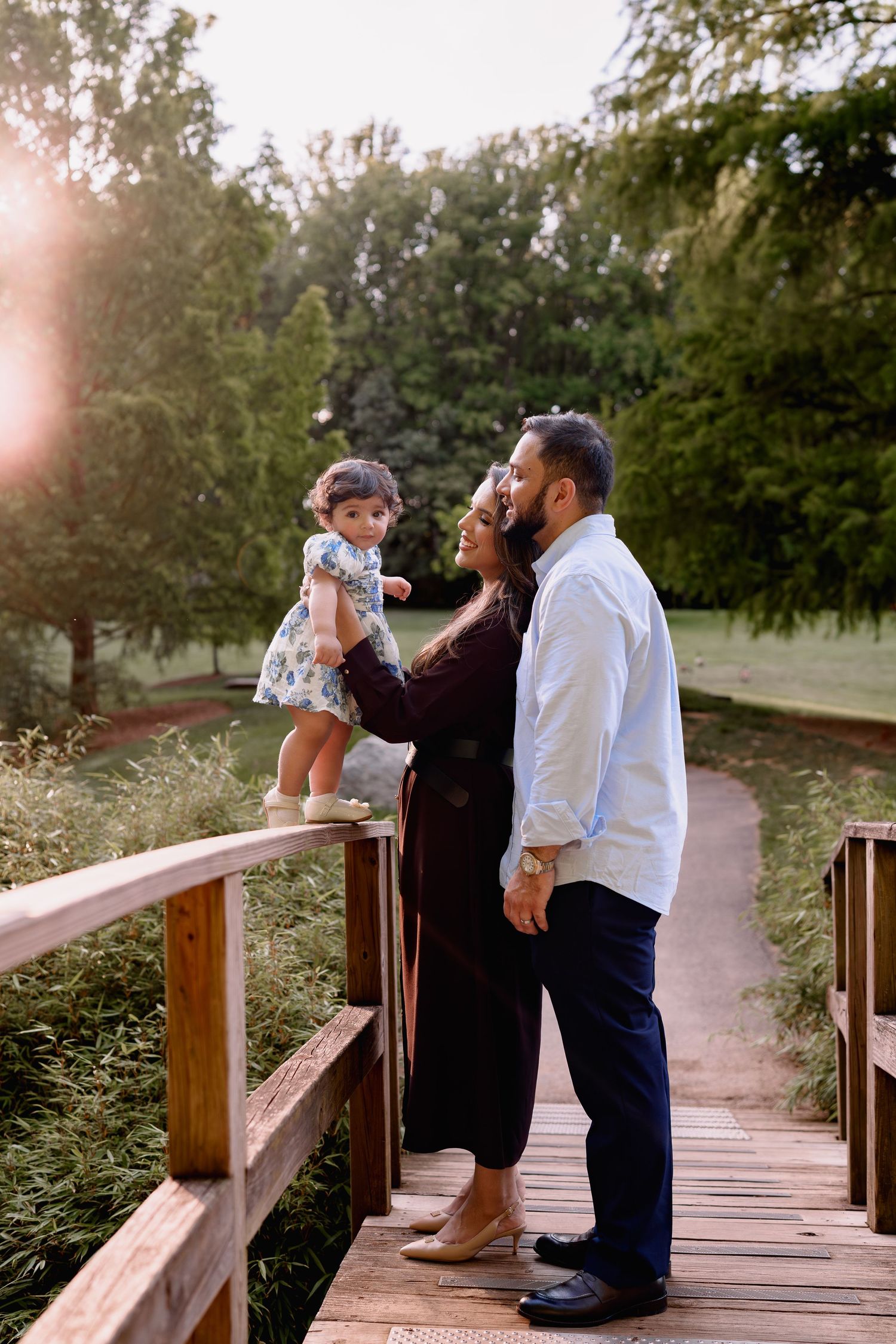 Family portrait on wooden bridge in park, parents kissing while holding toddler girl in floral dress at golden hour, in Wheaton, MD