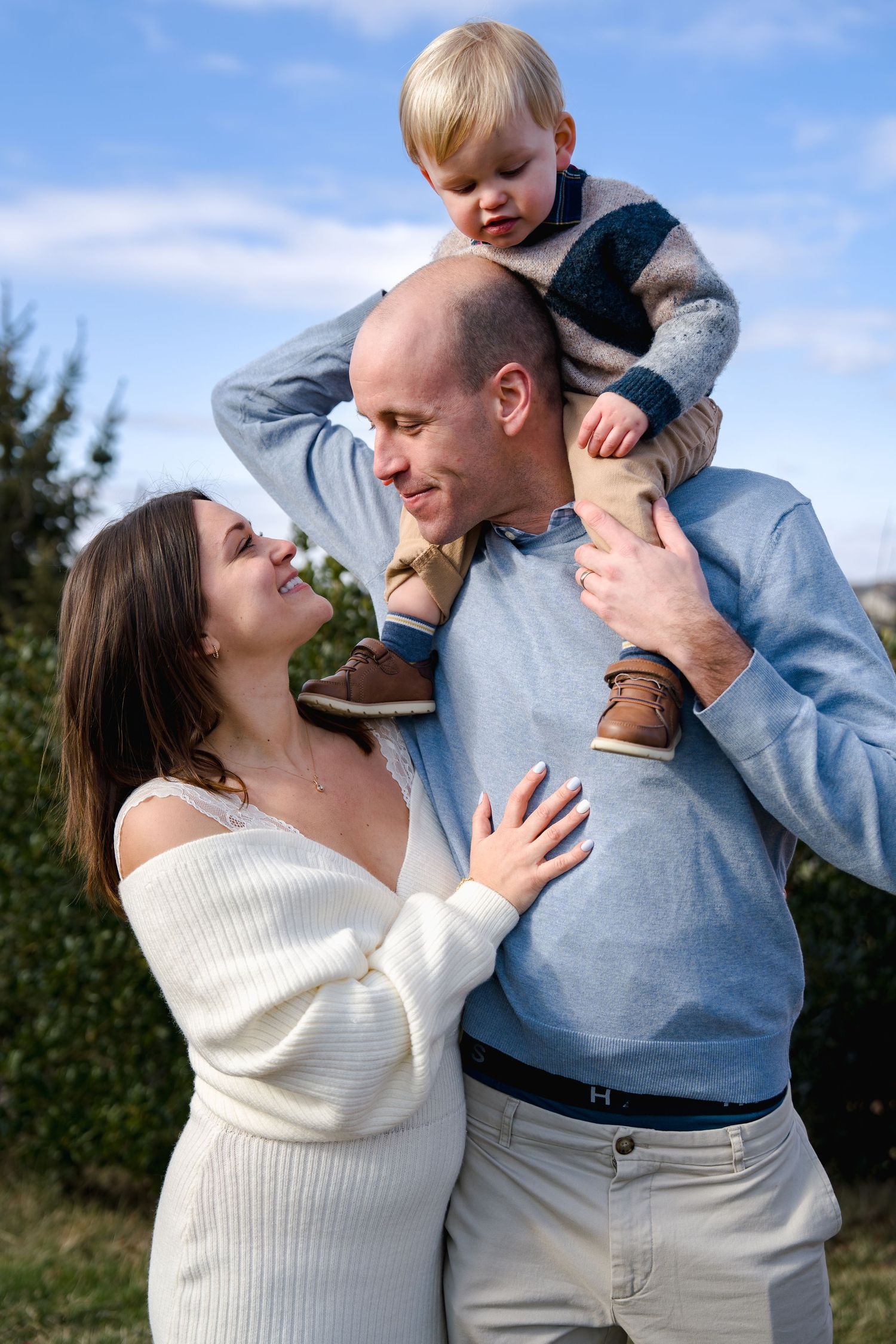 Happy family portrait outdoors, mother in white dress, father holding toddler on shoulders, blue sky background, in Frederick, MD