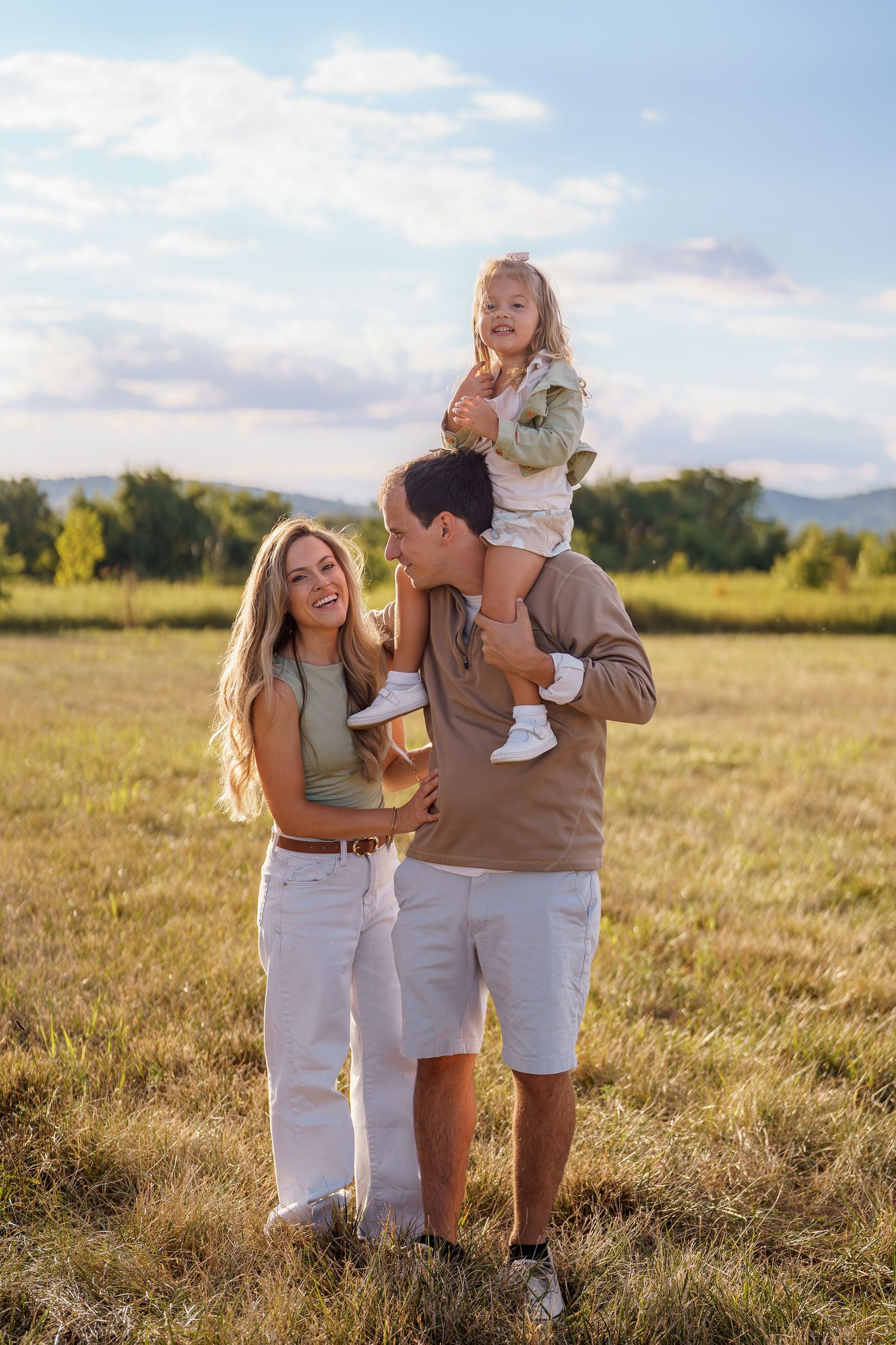 Happy family with young child on father's shoulders enjoying a sunny day in a green meadow with mountains in background, in Frederick, MD