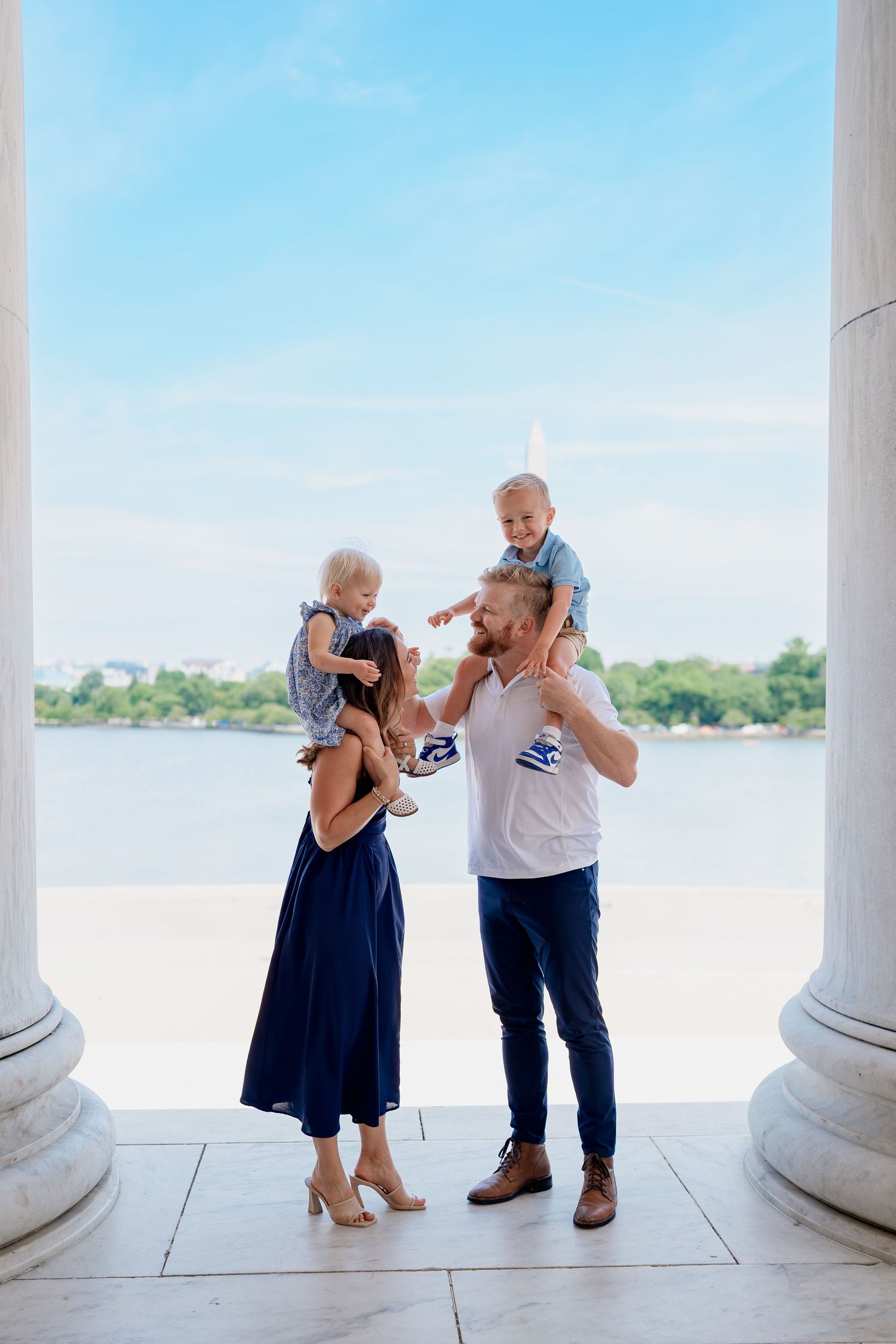 Family of four posing at Jefferson Memorial columns, parents holding toddlers with Potomac River view on sunny day.