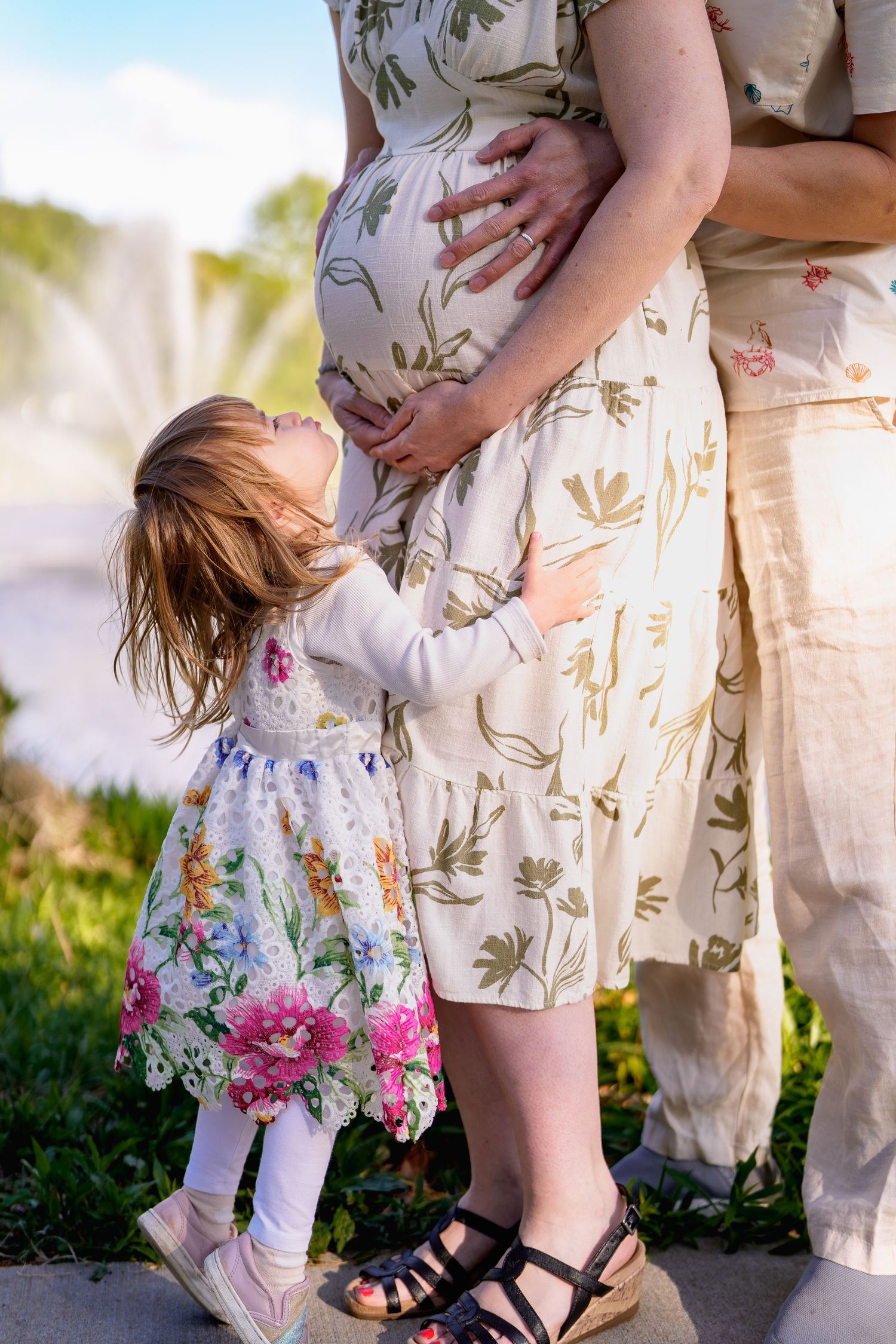 A young girl in a floral dress looks up at her pregnant mother's belly outdoors in a heartwarming maternity photo, in Vienna, VA