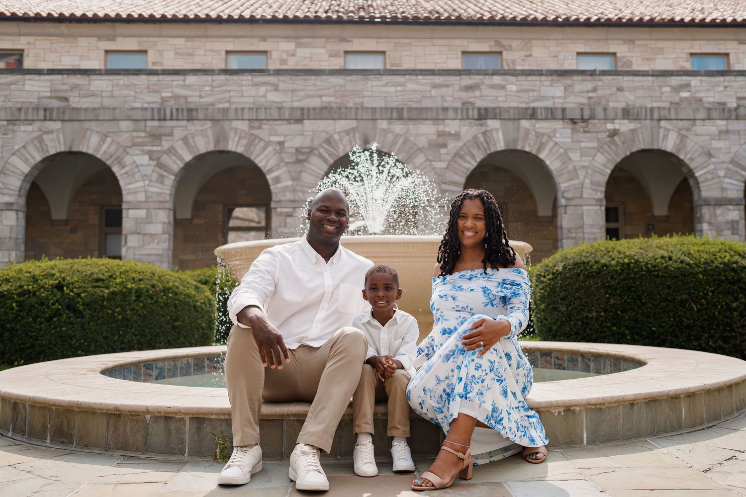 Happy family of three poses by a fountain courtyard, expecting mother in floral dress with husband and young son, in Ellicott City, MD
