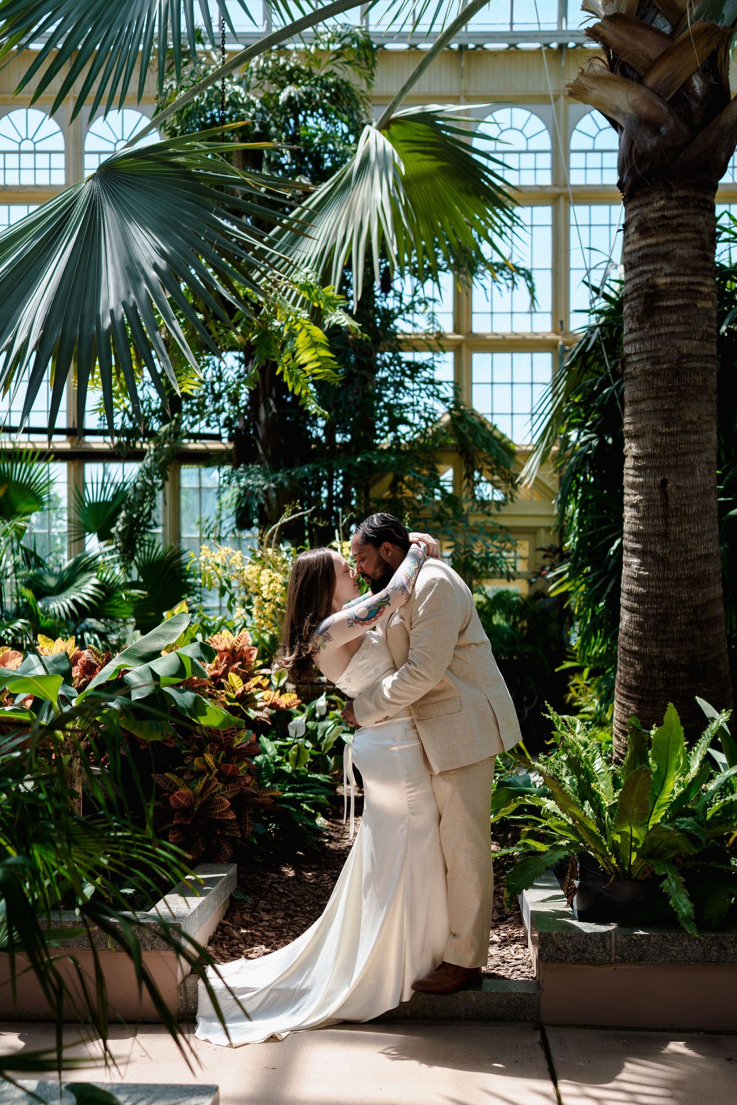 Bride and groom share a romantic embrace in a lush greenhouse conservatory, surrounded by tropical palms and colorful plants, in Maryland