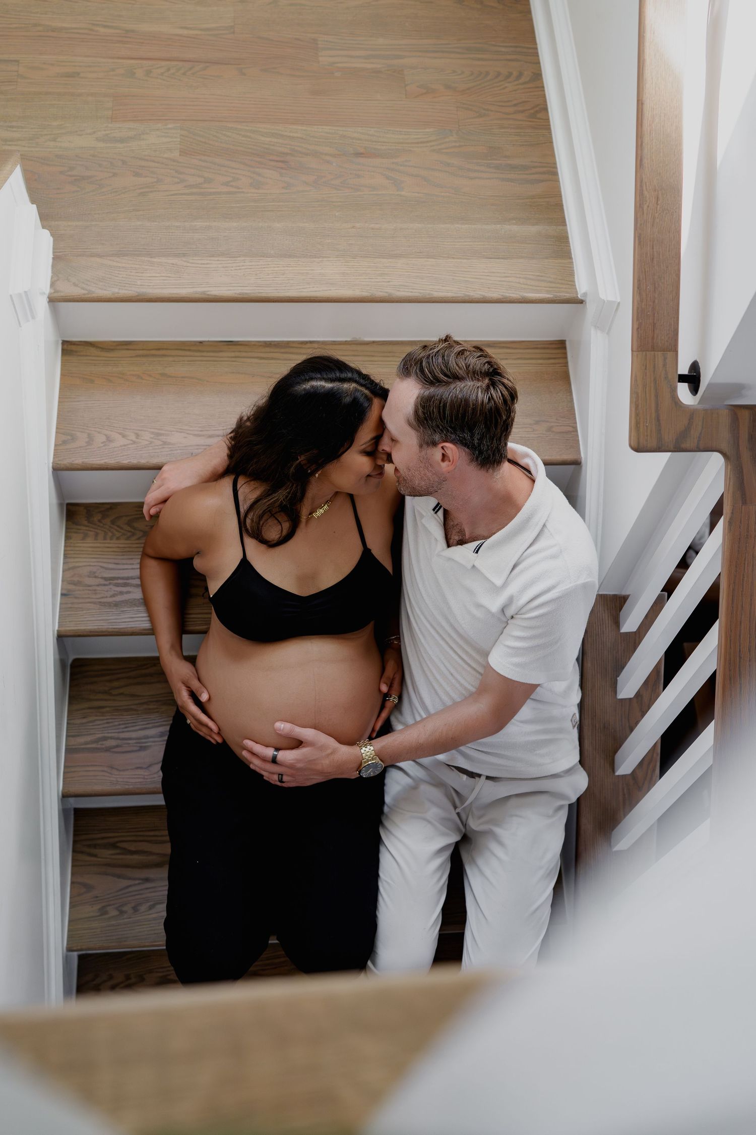 Couple sharing a tender kiss on wooden stairs, man gently holding pregnant woman's belly in a bright home, in Odenton, MD