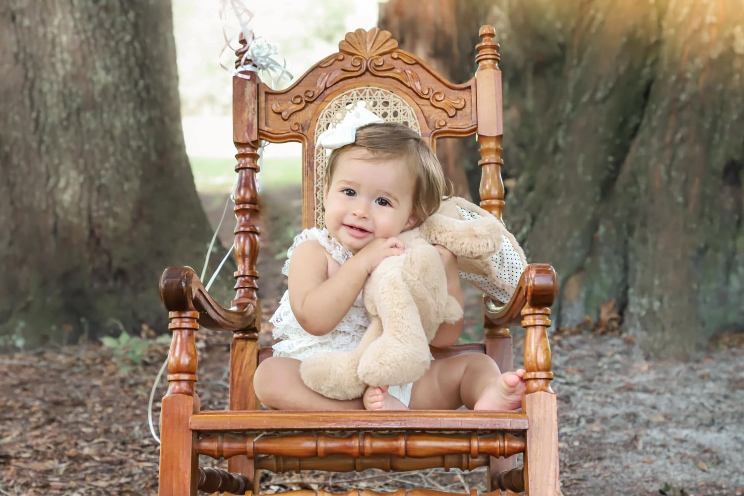 Adorable baby girl sitting in ornate wooden rocking chair outdoors hugging teddy bear wearing white outfit.